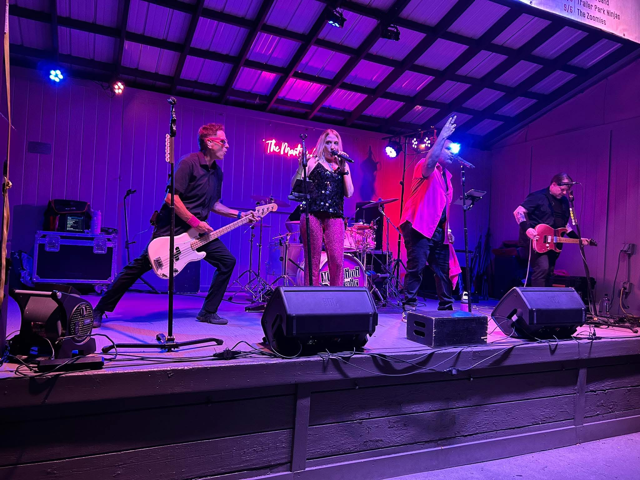 Four musicians perform on stage with purple and blue lighting, one woman singing, others playing guitars and drums.