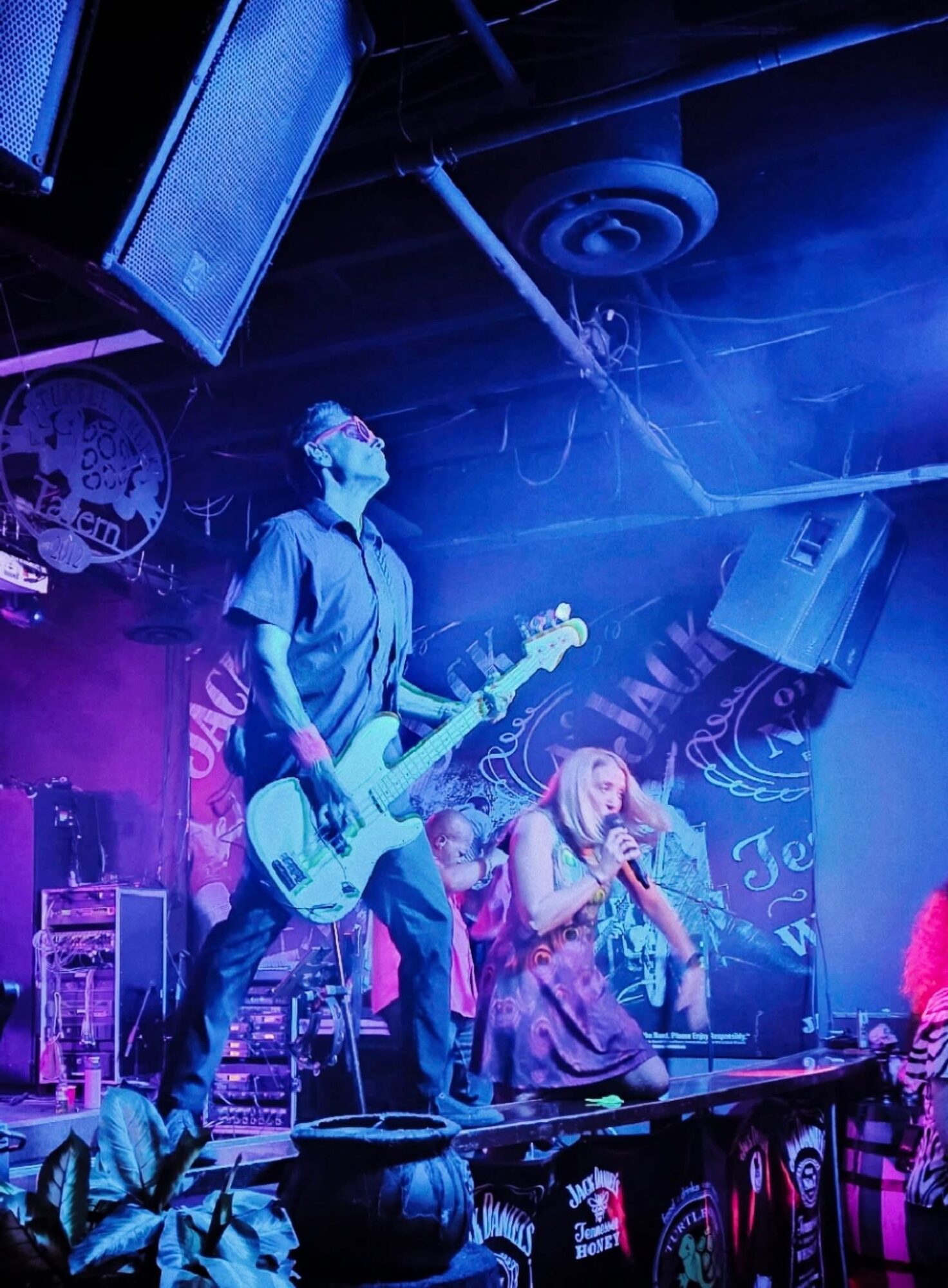 Musicians perform on stage with colorful lighting, one playing guitar and another singing into a microphone.
