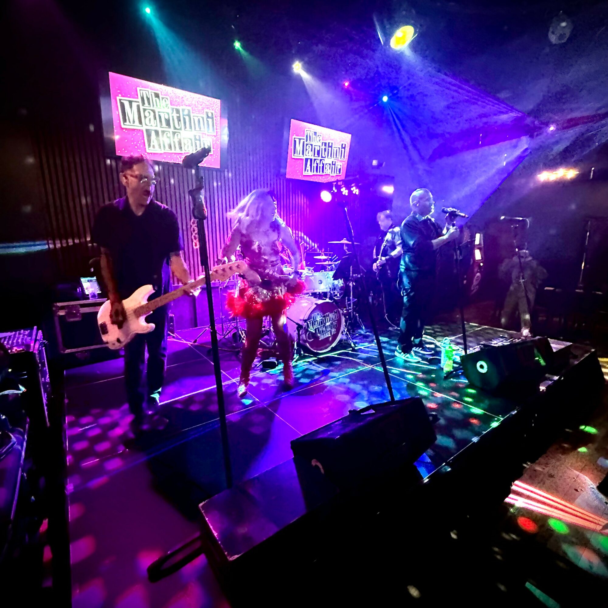 Band performing on stage with colorful lights and screens, musicians playing instruments, and microphones in front. Word signs are visible.