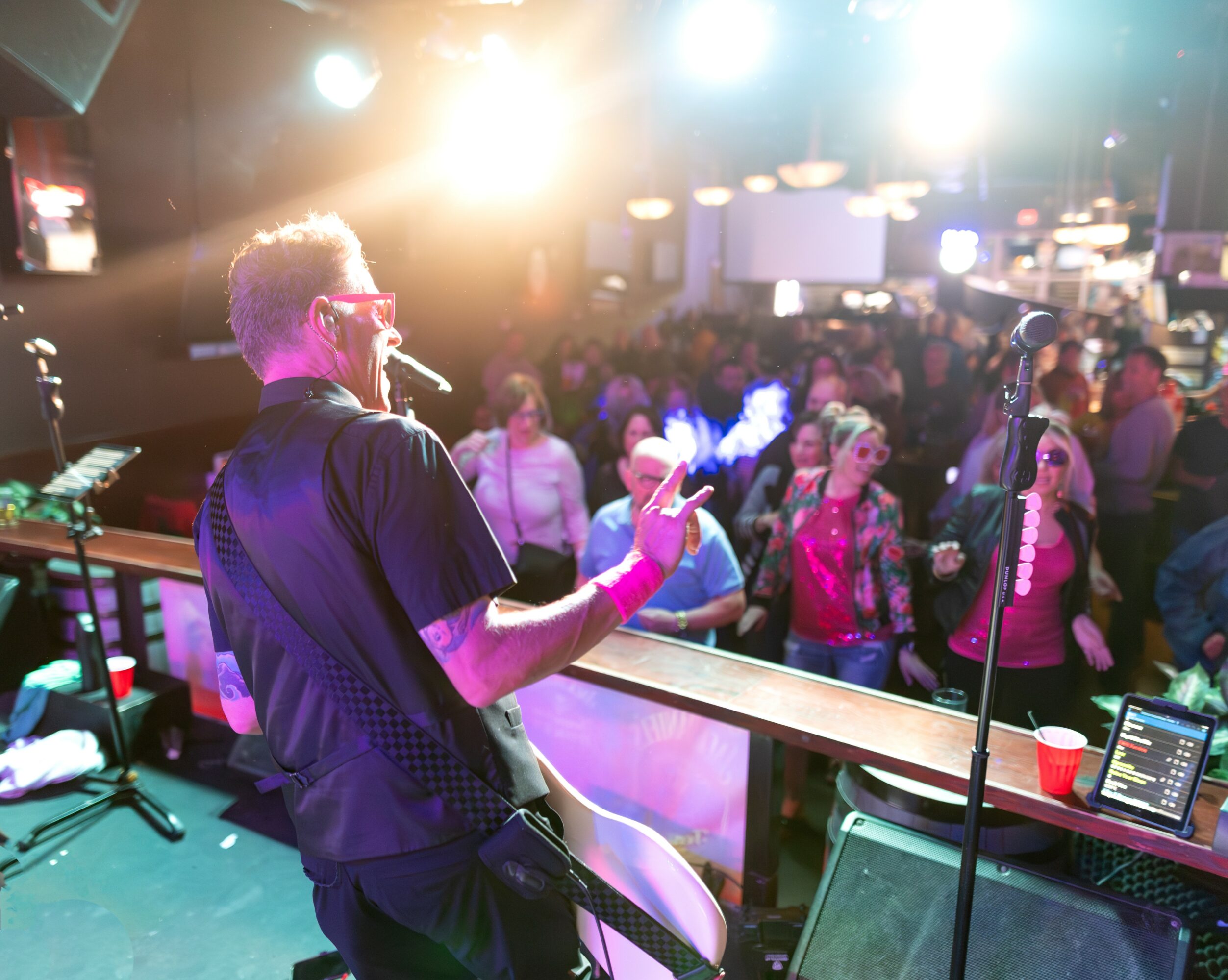 Musician performing on stage with a crowd dancing in a lively indoor venue, bright stage lights overhead.