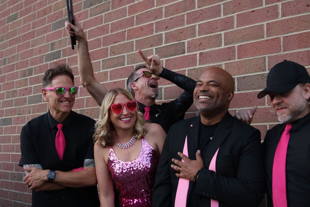 Group of five people smiling and posing against a brick wall, some wearing pink accessories and sunglasses.