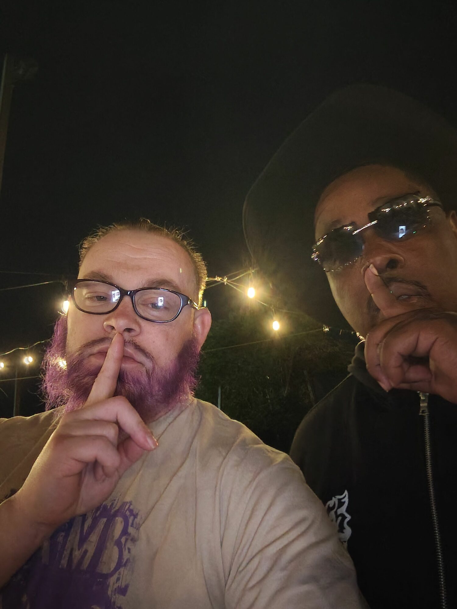 Two people, one with glasses and purple hair, making a gesture near their face, outdoors at night with string lights.