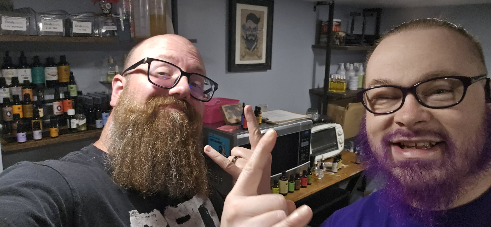 Two men with glasses and beards smiling and making a peace sign in a room with shelves and equipment