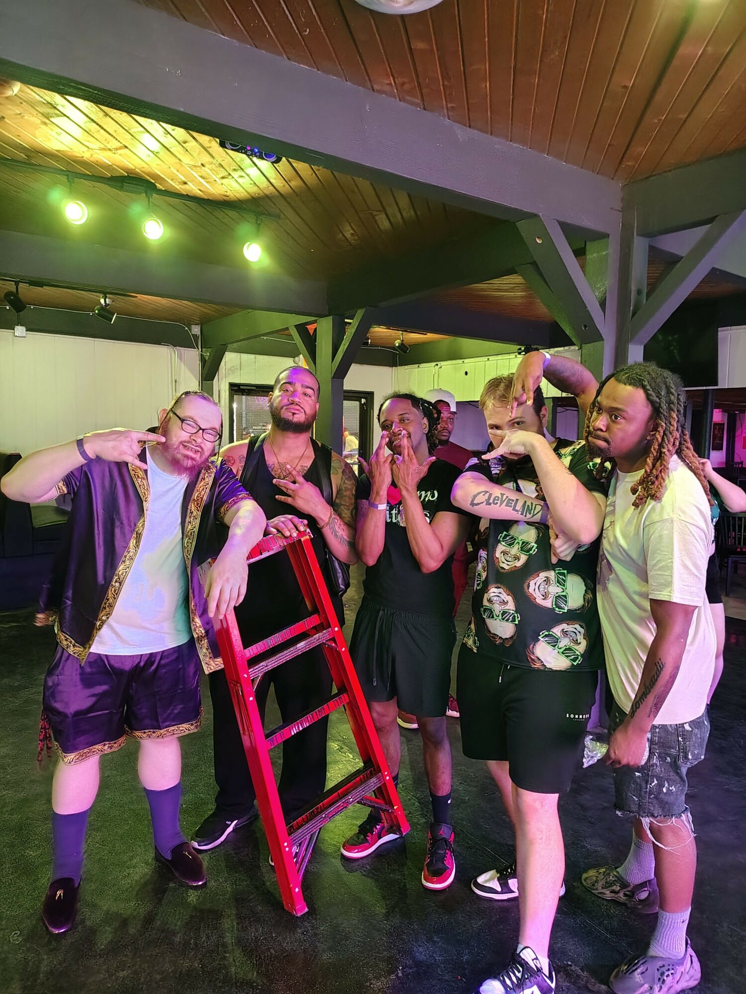 Group of people standing indoors, some holding a red chair, with a wooden ceiling and green lighting in the background.