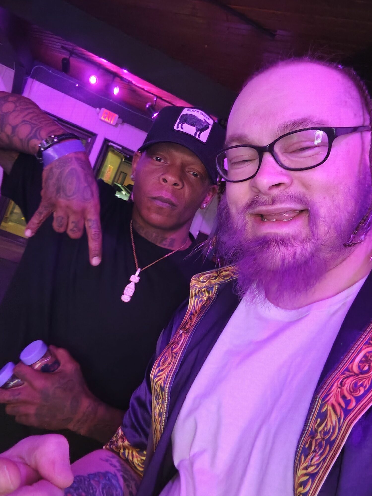 Two men, one with glasses and a cap, smiling, in a dimly lit environment with purple lighting.