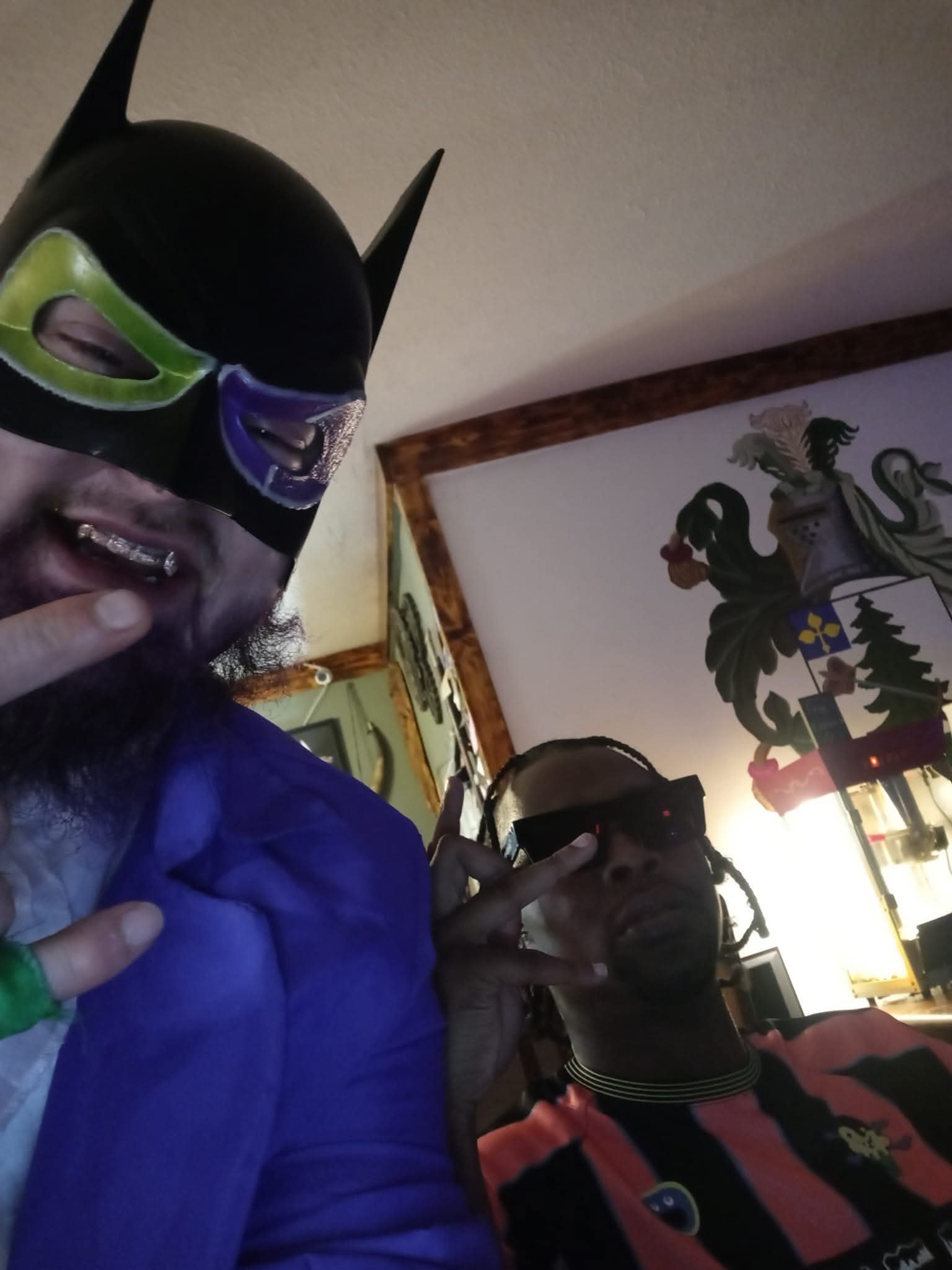 Two people wearing masks and costumes, one with a Batman mask and the other with sunglasses, indoors with a decorative wall.