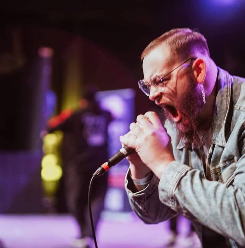 Man singing passionately into a microphone on stage with colorful lights in the background.
