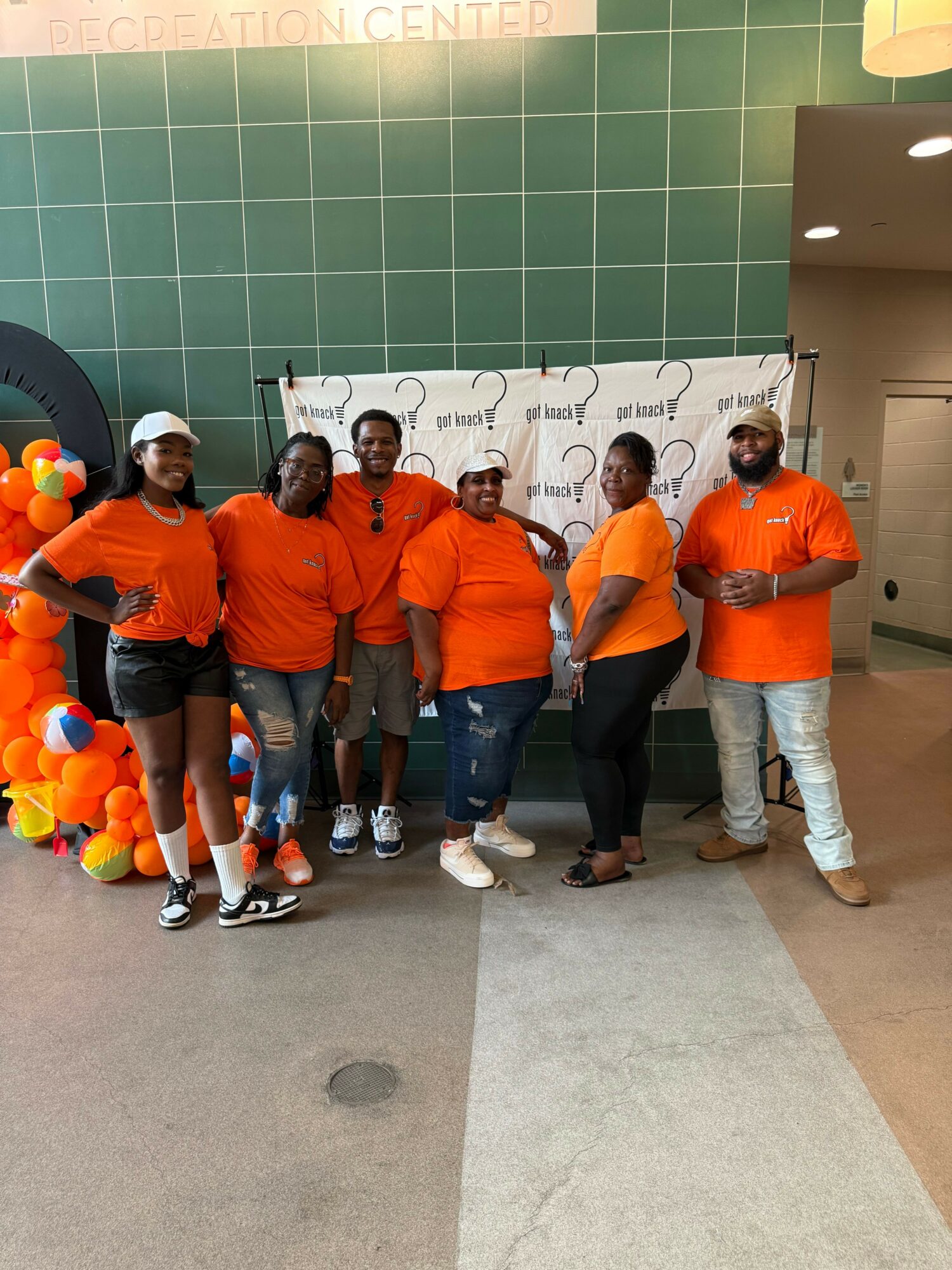 Group of six people wearing orange shirts standing in front of a white backdrop with a logo, indoors. Some are holding balloons.