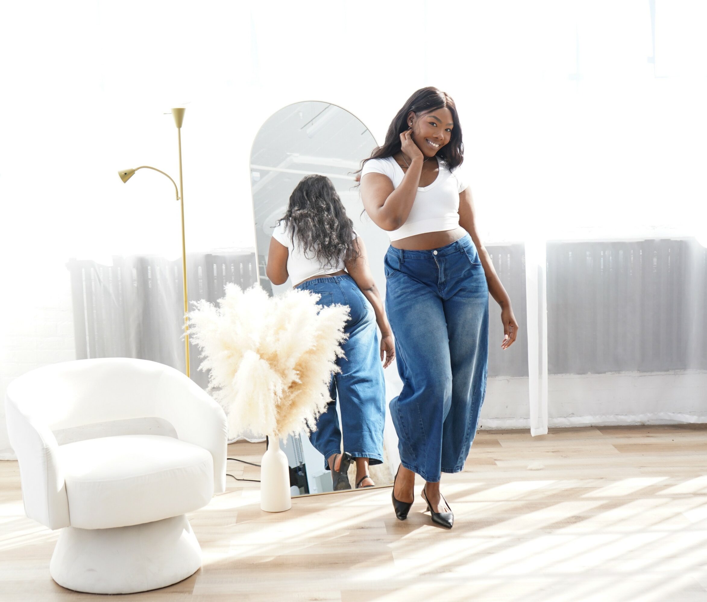 Two women in a bright room, one looking in a mirror, the other walking, with a white chair and a tall lamp.