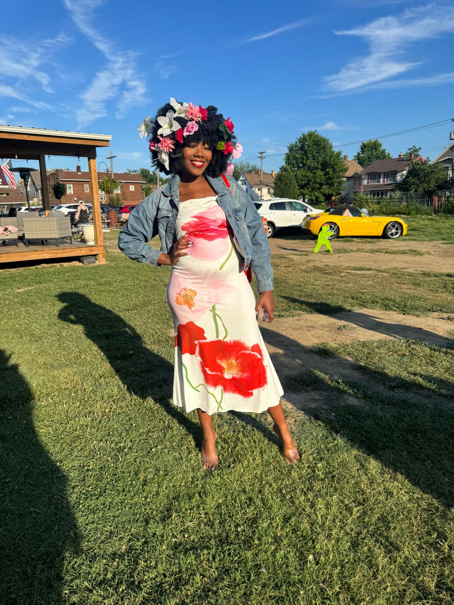 Woman in a floral dress and flower crown walking on grass, with a yellow vehicle and a building in the background.