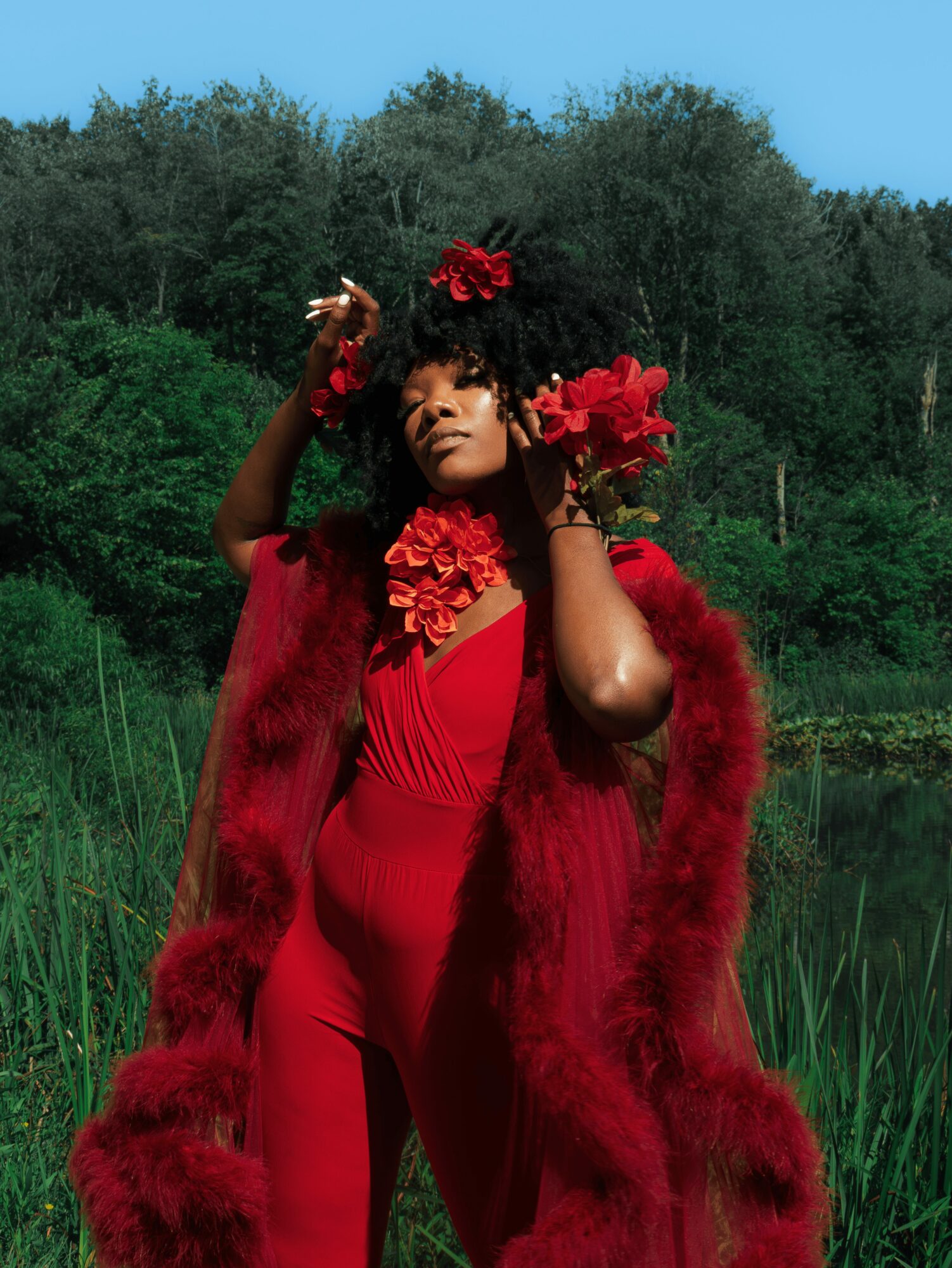 Woman in red dress and fur coat outdoors, holding red flowers in her hair, with trees and blue sky behind her.
