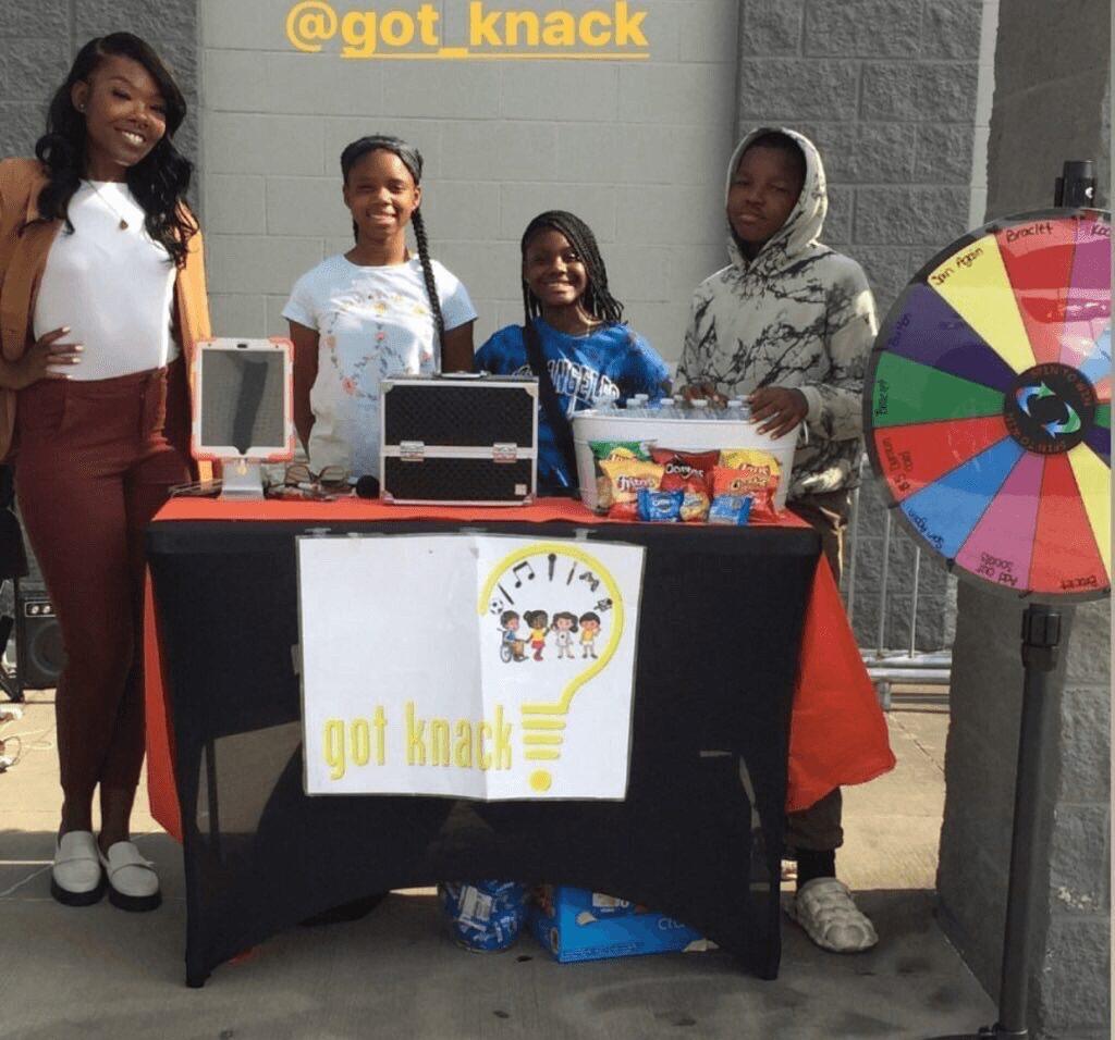 Four women stand behind a table with a sign, a prize wheel, and various items, outdoors on a sidewalk.
