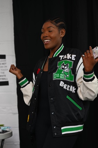 Young woman smiling, wearing a black and green varsity jacket with patches, standing indoors against a dark background.