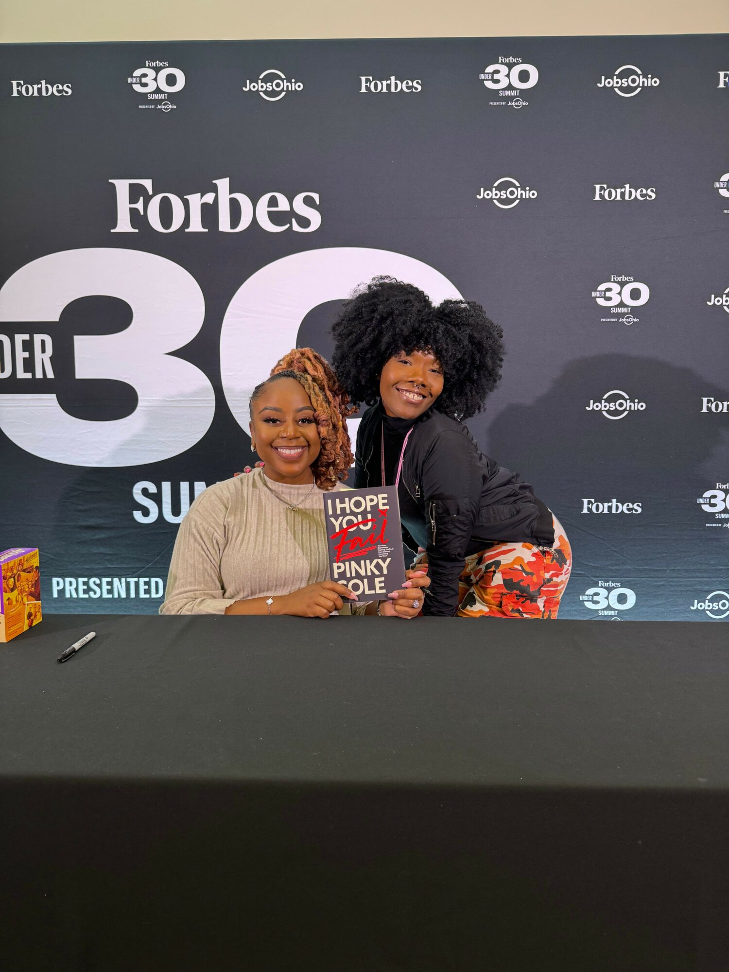 Two women sitting at a table with a black background and Forbes logo, smiling for the camera.