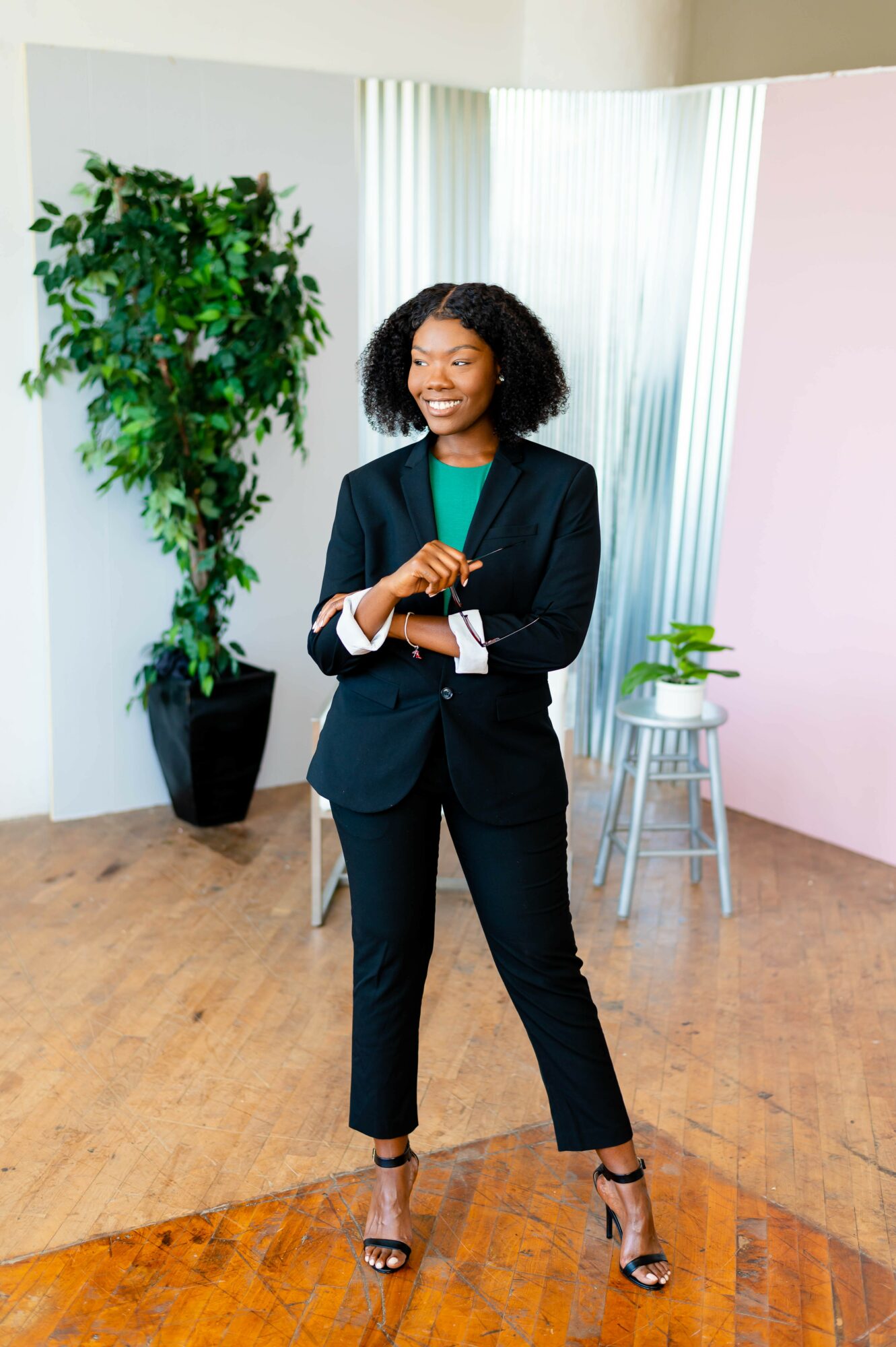Woman in a dark suit standing with arms crossed, smiling, in a room with plants and curtains.