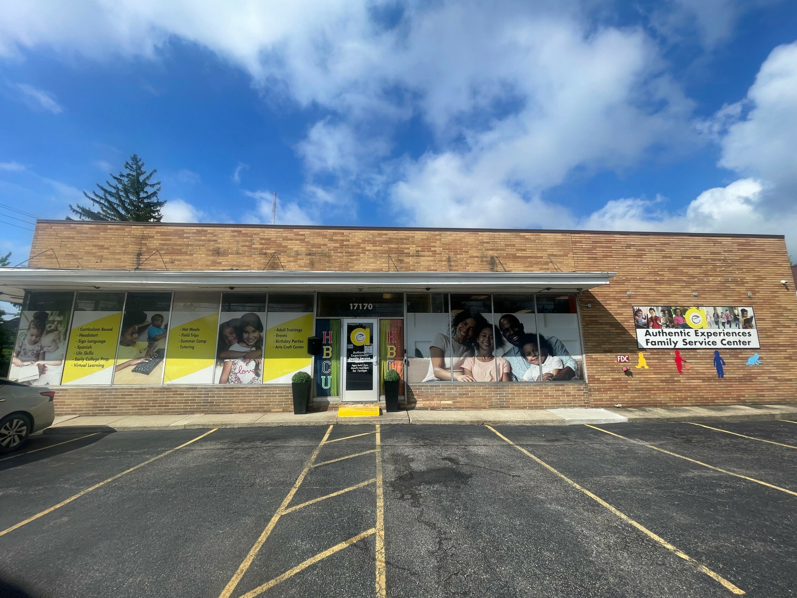 Single-story building with large windows, parking lot in front, blue sky with clouds, and a sign on the right side.