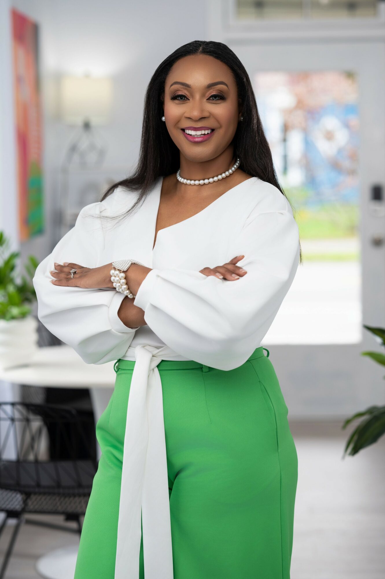 Smiling woman with crossed arms, wearing a white blouse and green pants, standing indoors with a bright background.