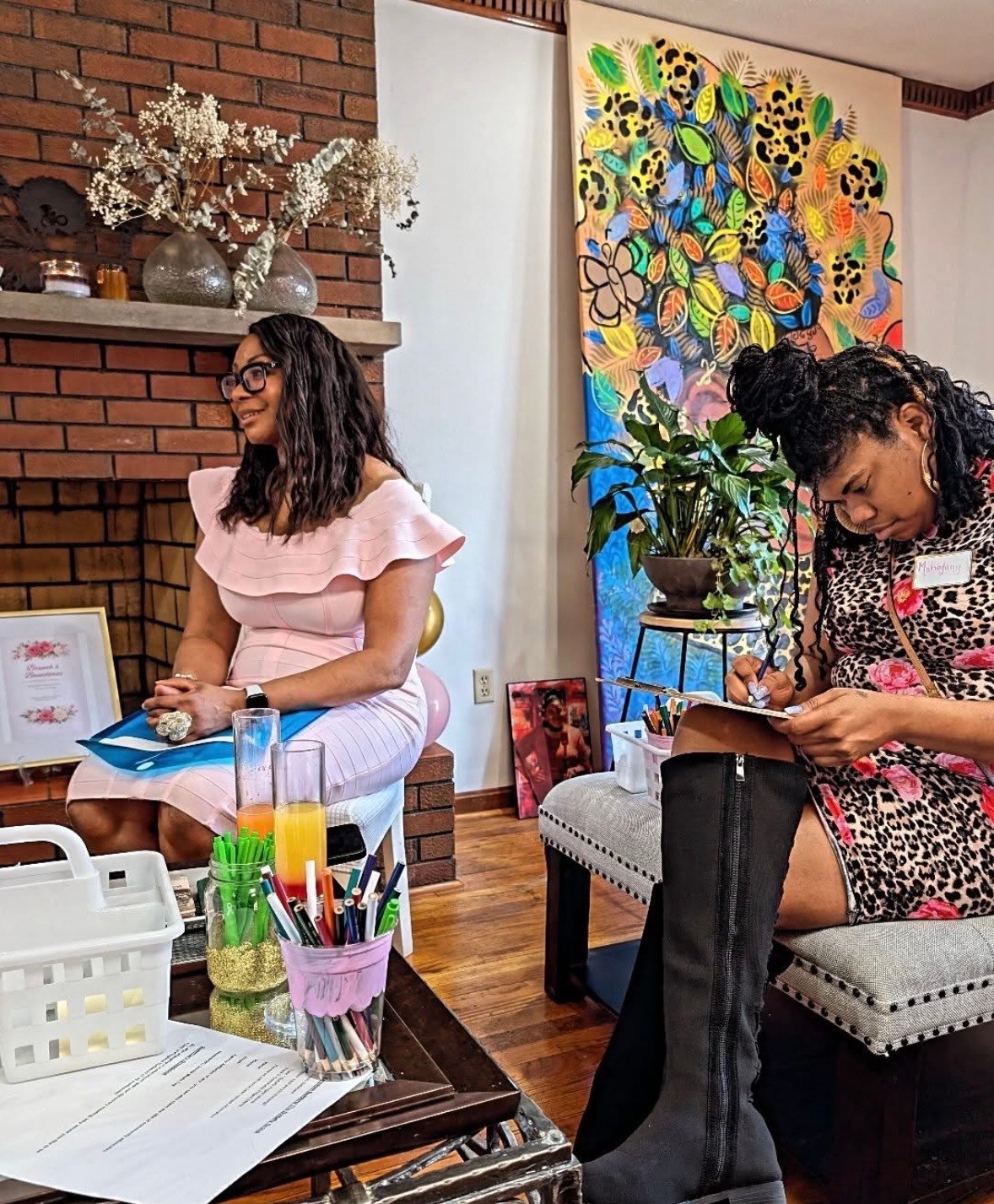 Two women sitting indoors, one reading a book and the other smiling, with colorful artwork and plants in the background.