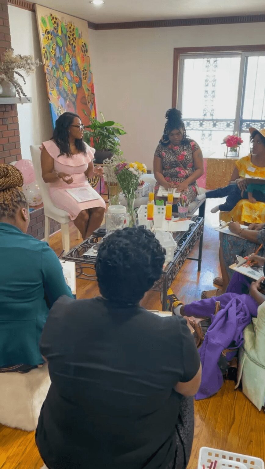 Group of women sitting around a table in a room with large window, colorful artwork, and plants, engaged in discussion.
