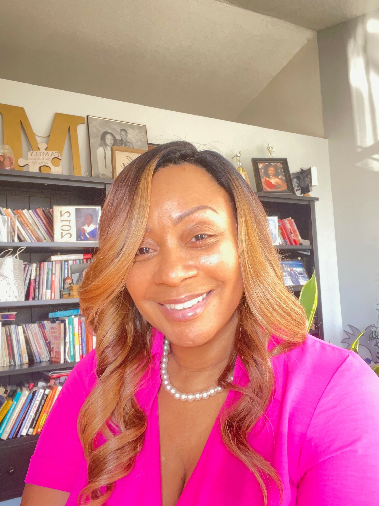Woman with long wavy hair wearing a pink top and pearl necklace, smiling indoors with bookshelves in background.