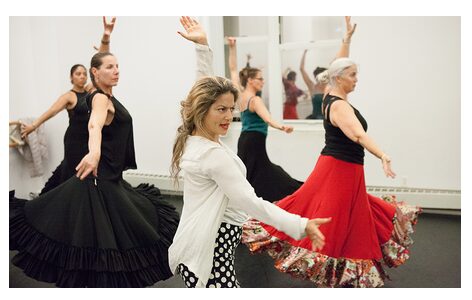 Group of women dancing in a studio, some wearing long skirts, one in a white jacket, others in black and red skirts.