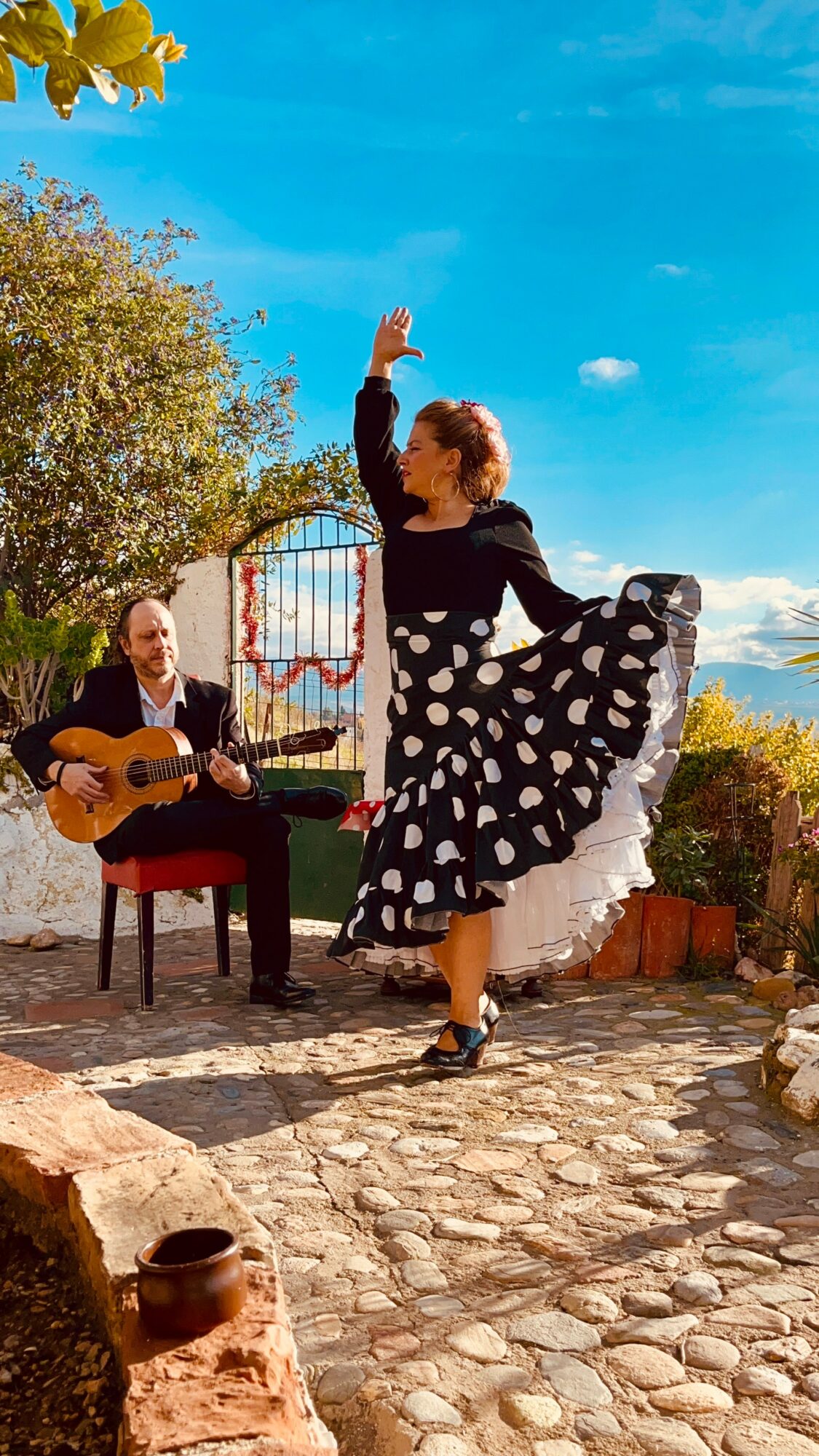 Woman dancing outdoors with a man playing guitar, trees and blue sky in background.