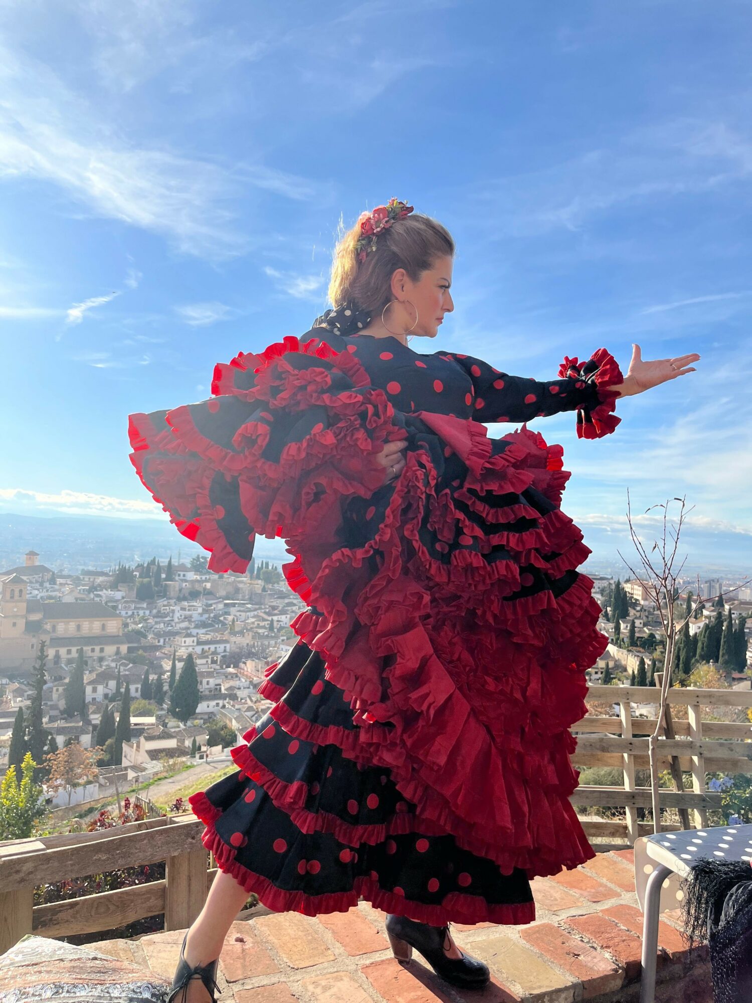 Woman in a red and black dress with floral details standing outdoors on a brick surface, with a cityscape and blue sky in background.