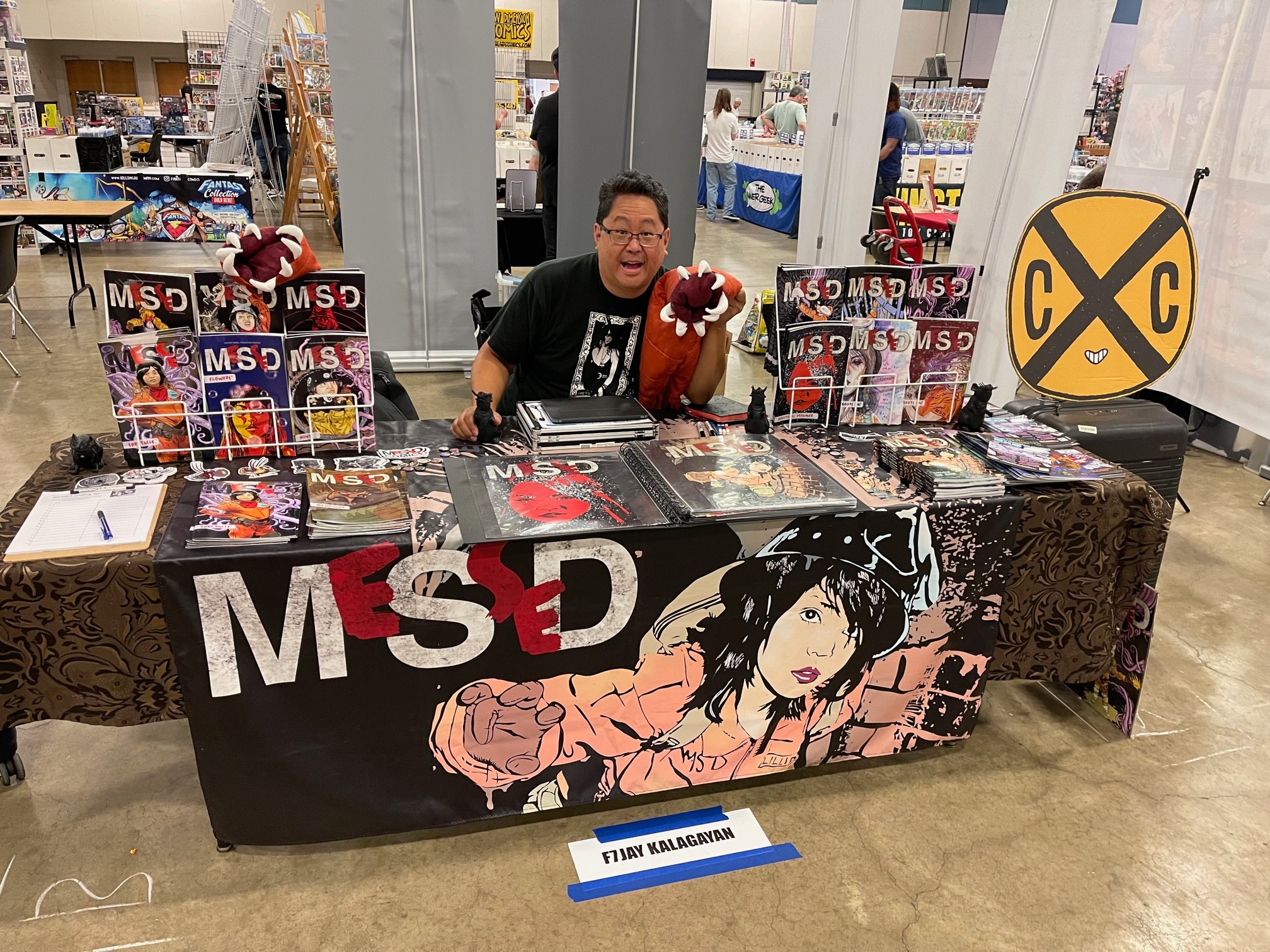 Person sitting behind a table with manga and merchandise, store shelves in background, large MS-D logo on tablecloth, and a round sign with crossed-out X and C.