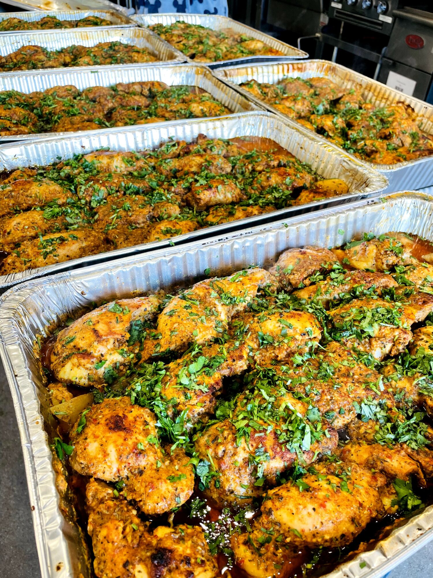 Multiple aluminum trays filled with cooked, seasoned chicken pieces garnished with herbs, arranged on a countertop.