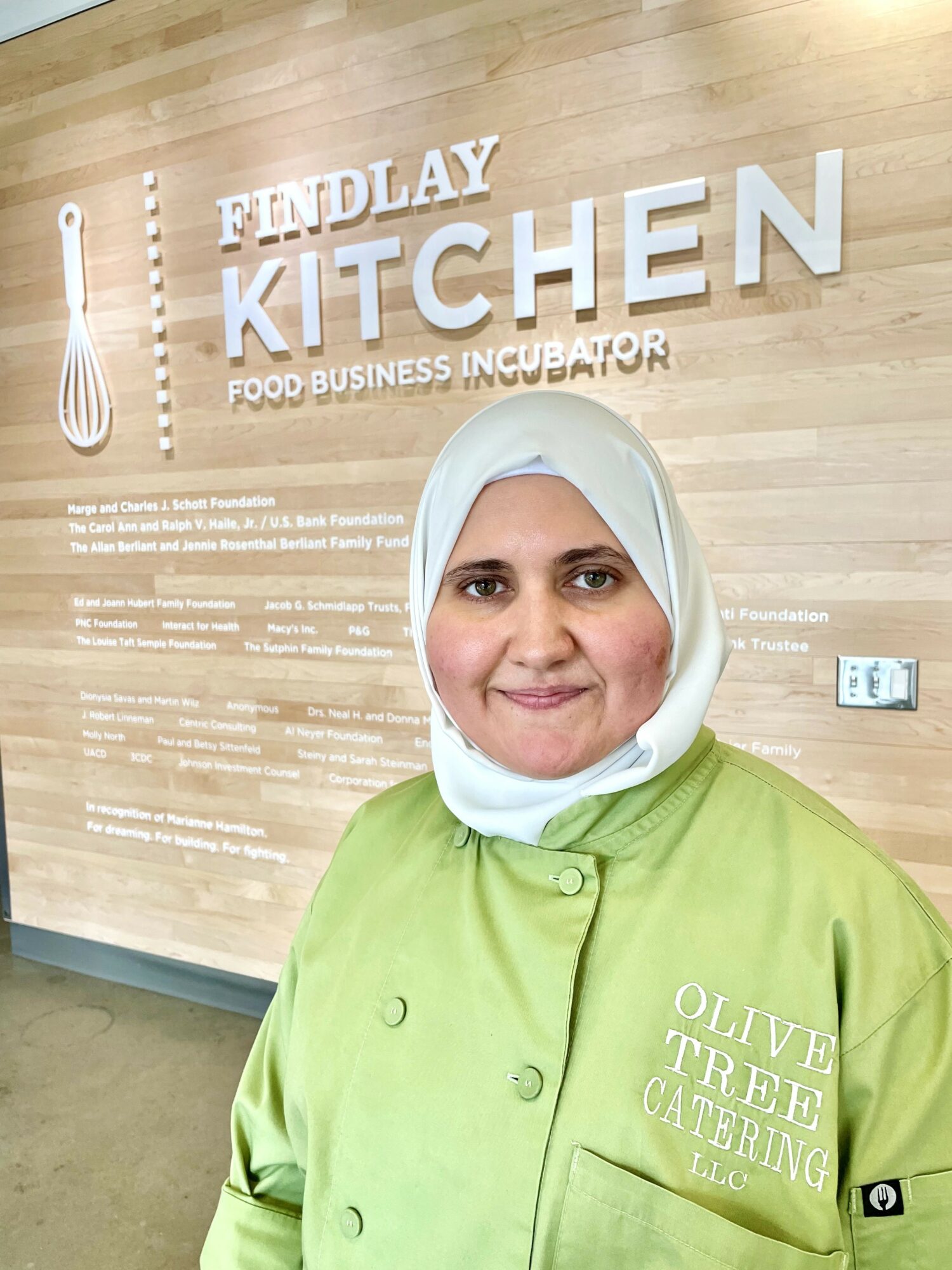 Woman in green chef's coat and white hijab standing in front of a wooden wall with 'Findlay Kitchen Food Business Incubator' sign.