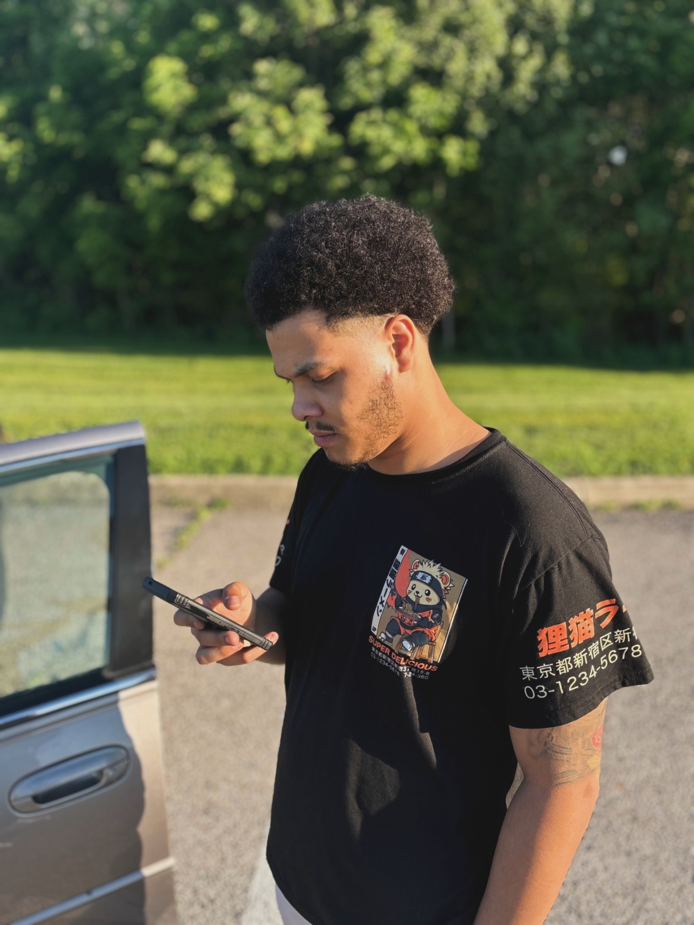 Young man with curly hair looking at his phone outdoors, standing next to a vehicle, with trees in the background.