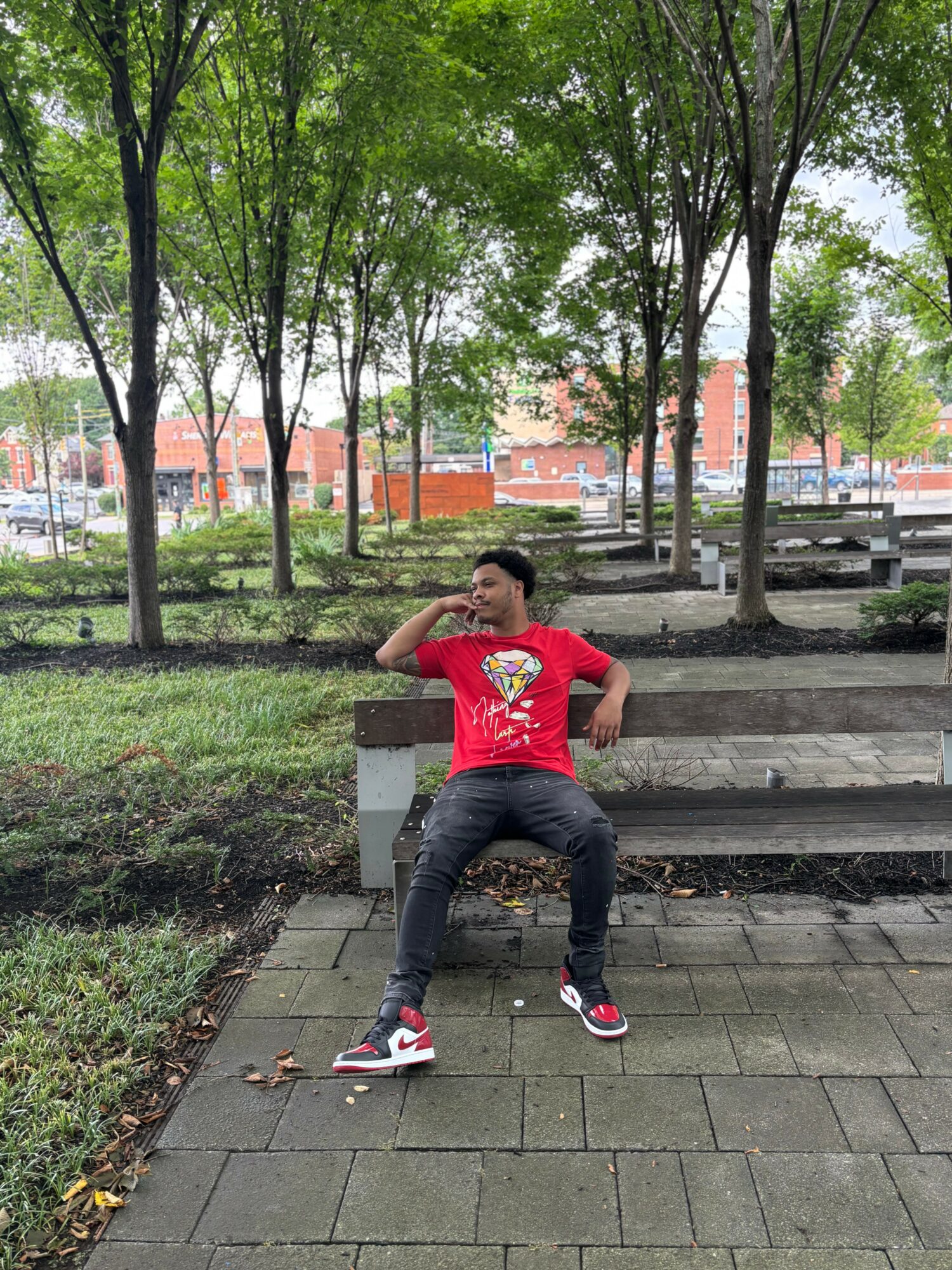 Person sitting on steps in park, wearing red shirt and black pants, surrounded by trees and benches.