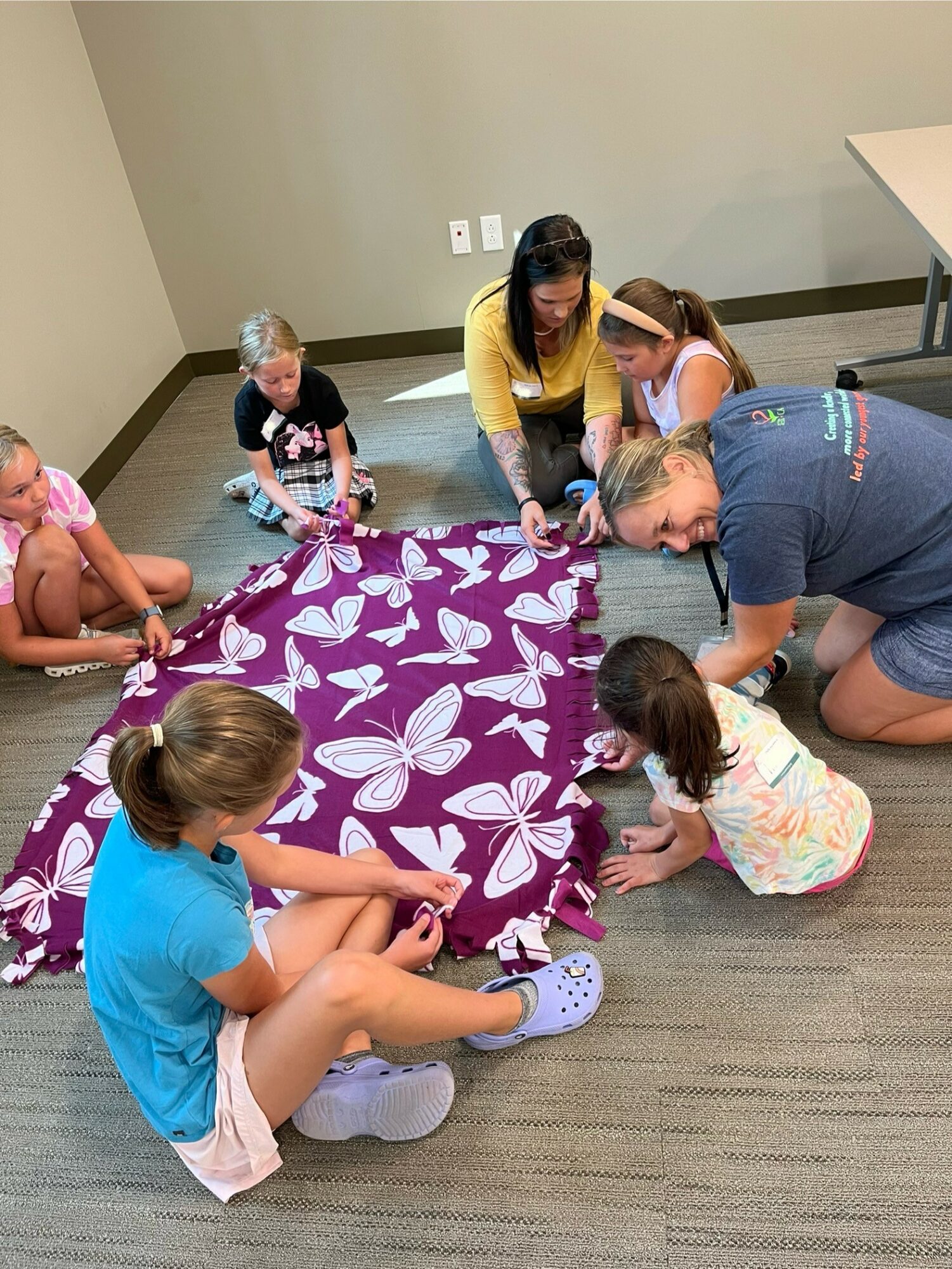 Group of children and adults gathered around a purple cloth with white butterfly patterns on a carpeted floor, engaging in an activity.