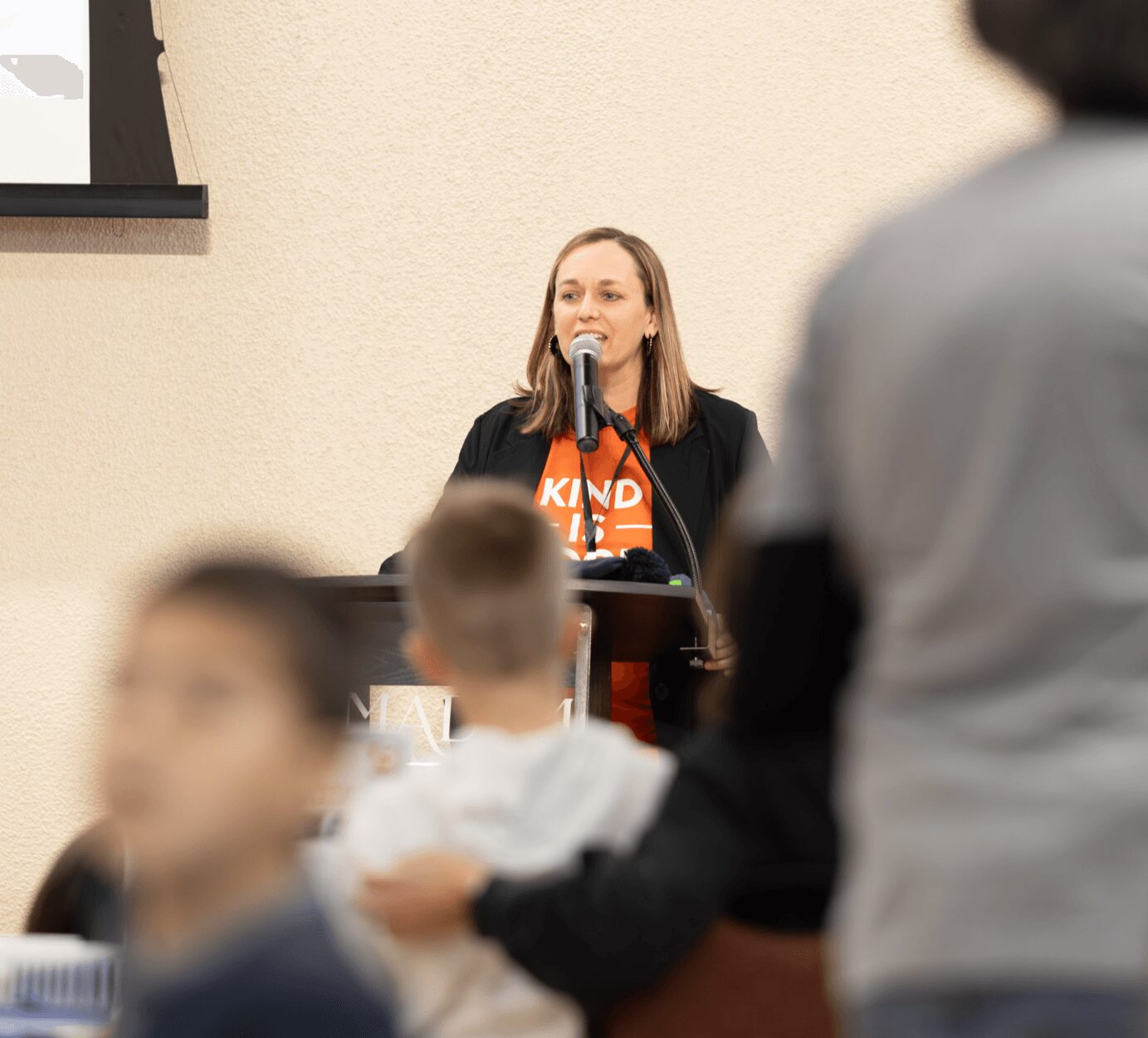 Woman speaking at a podium with a microphone, wearing a black blazer and orange shirt, in a room with beige walls.