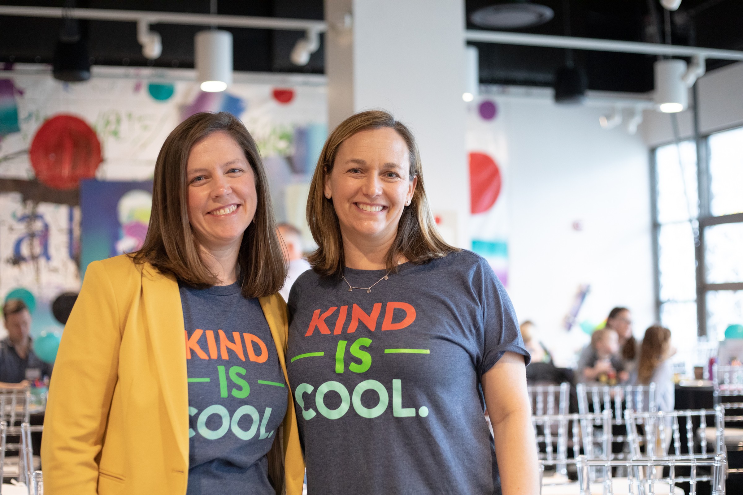Two women smiling, one wearing a yellow blazer and the other a navy T-shirt with colorful text, in a bright indoor space.
