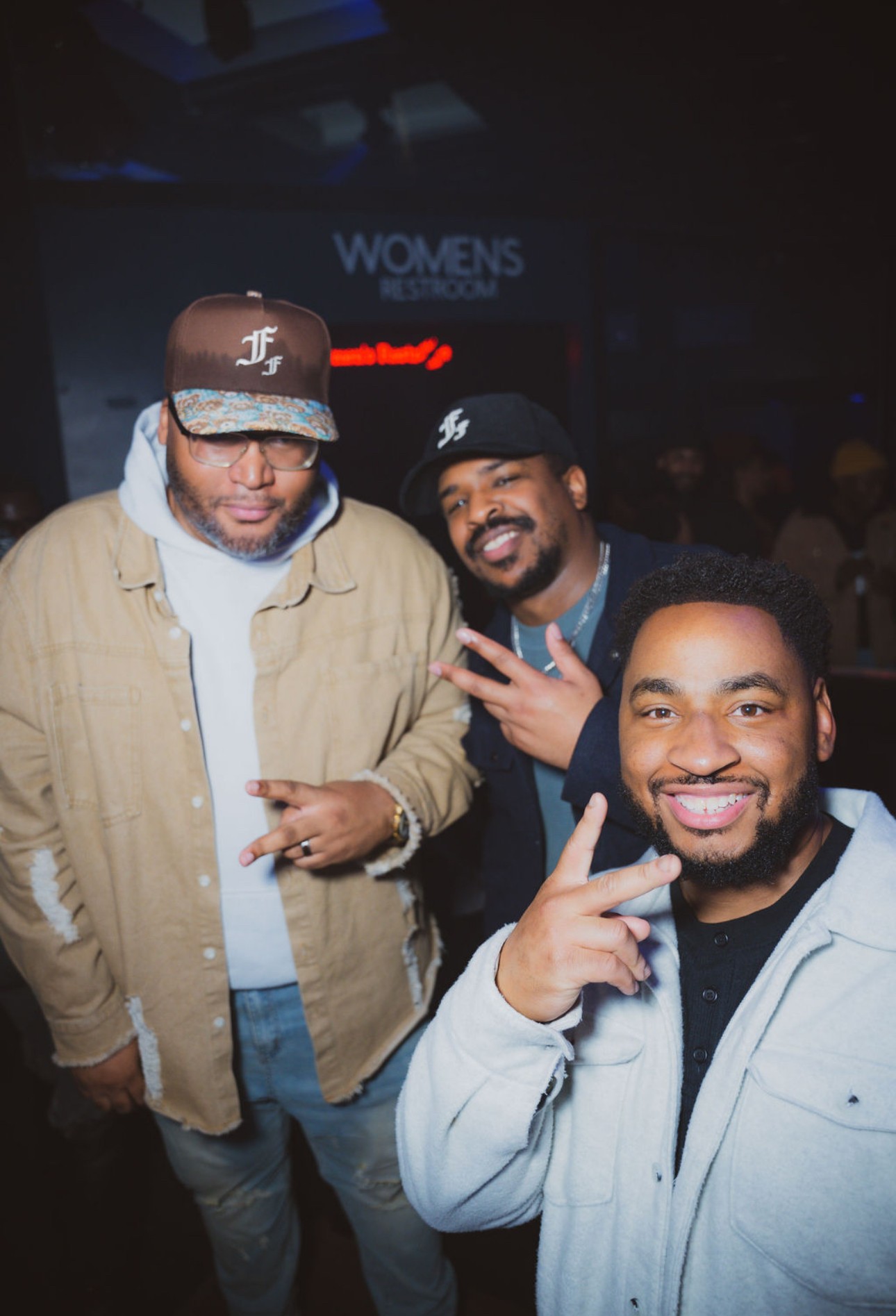 Three men smiling and posing for a photo in a dark indoor setting, with a sign reading 'WOMENS' in the background.