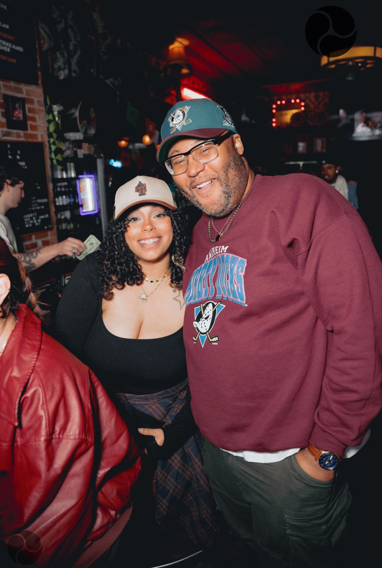 Two people smiling in a dimly lit bar, one wearing a baseball cap and glasses, the other with curly hair and a hat.