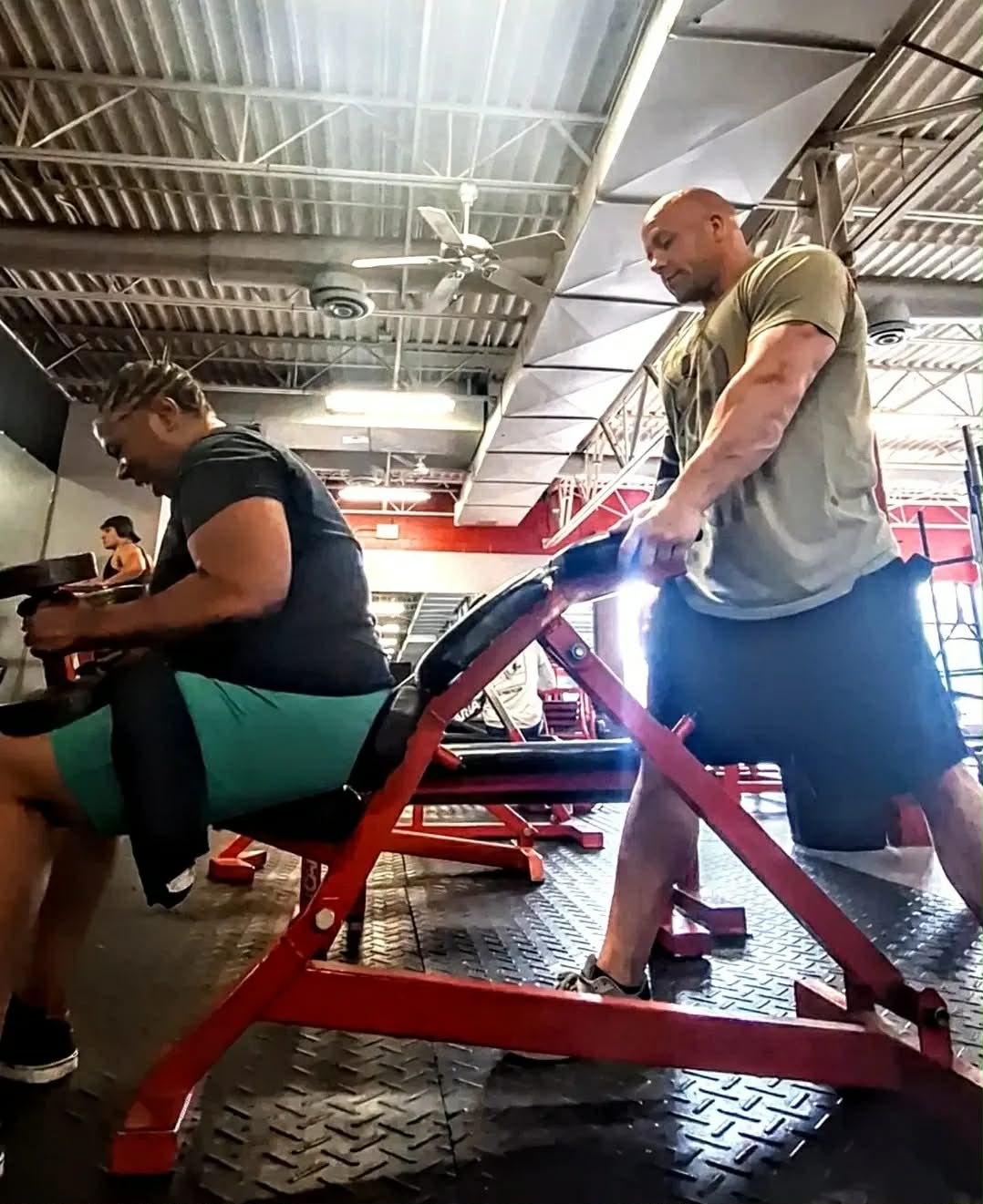 A man is kneeling on a red workout machine in a gym, assisting a person sitting on it. The gym has a high ceiling with metal beams and bright lighting.
