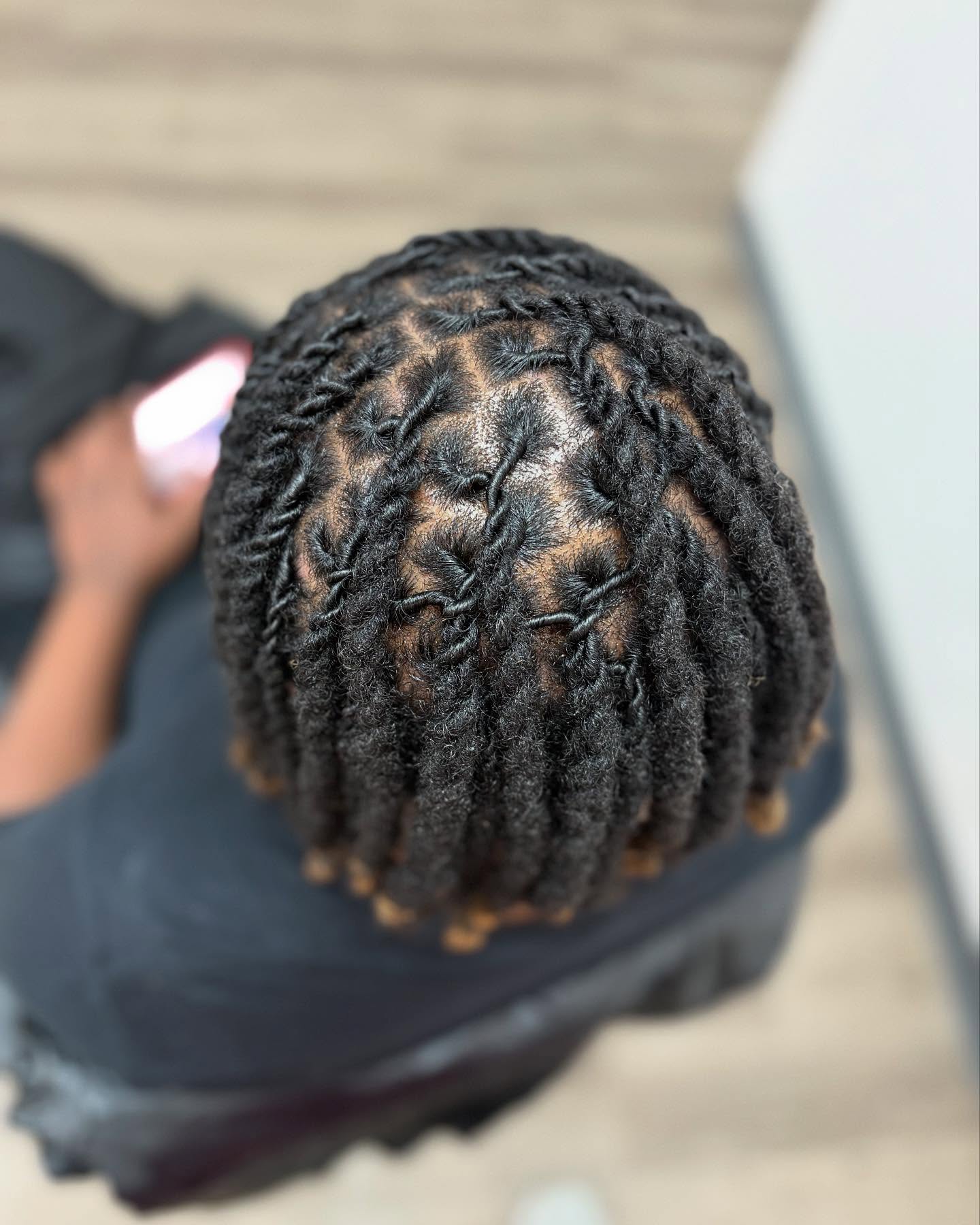 Top view of a person with neatly styled, thick, twisted dreadlocks on their head, sitting indoors on a wooden floor.