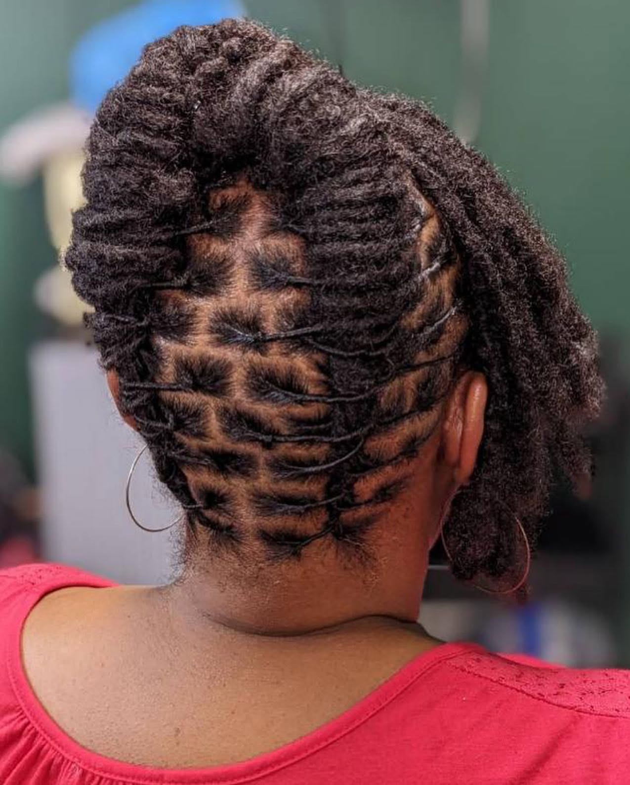 Back of a person's head with styled natural hair in twists and locs, wearing a red top.