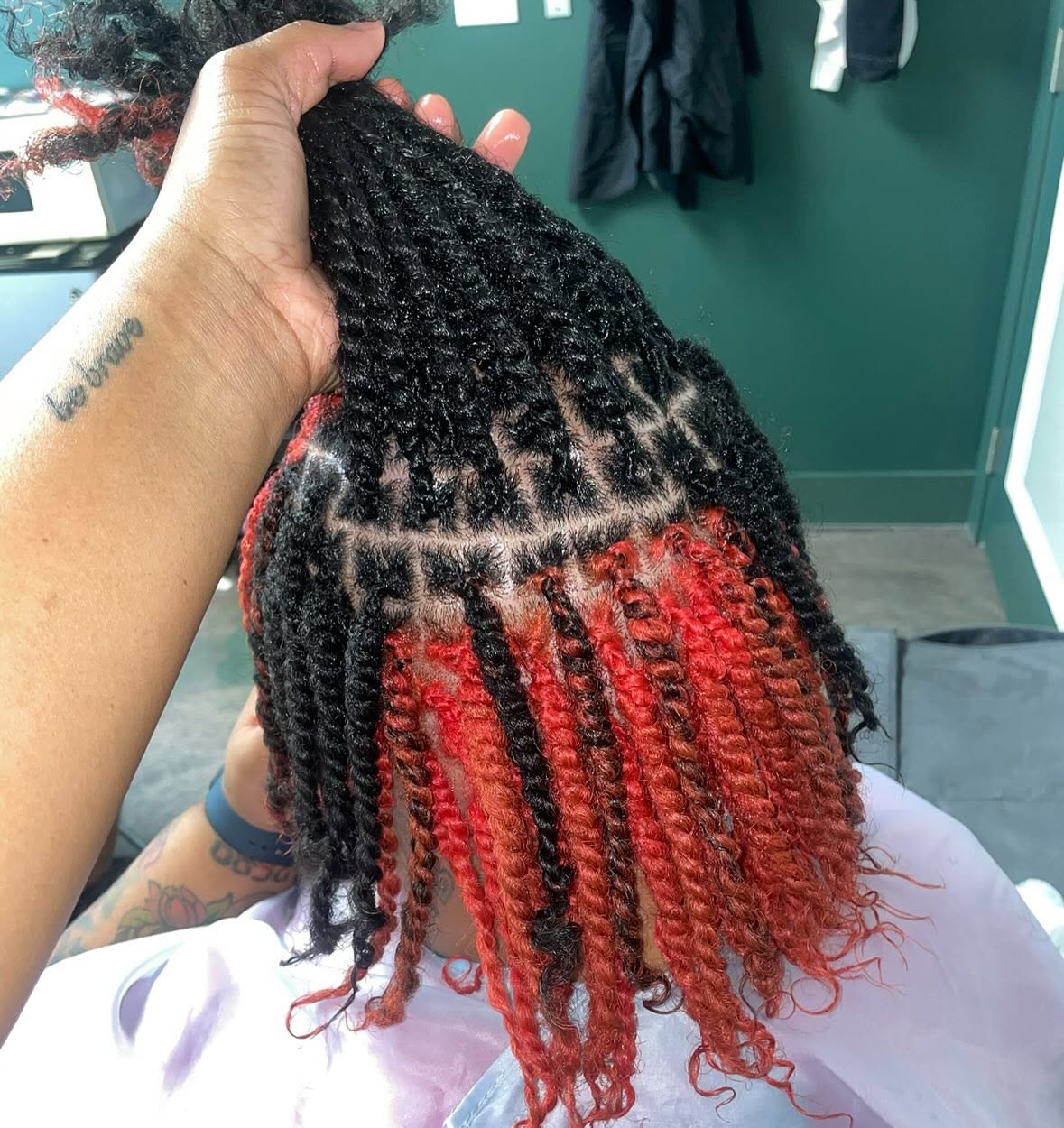 Person holding styled hair with black roots and red tips, in a salon setting, with a green wall in the background.