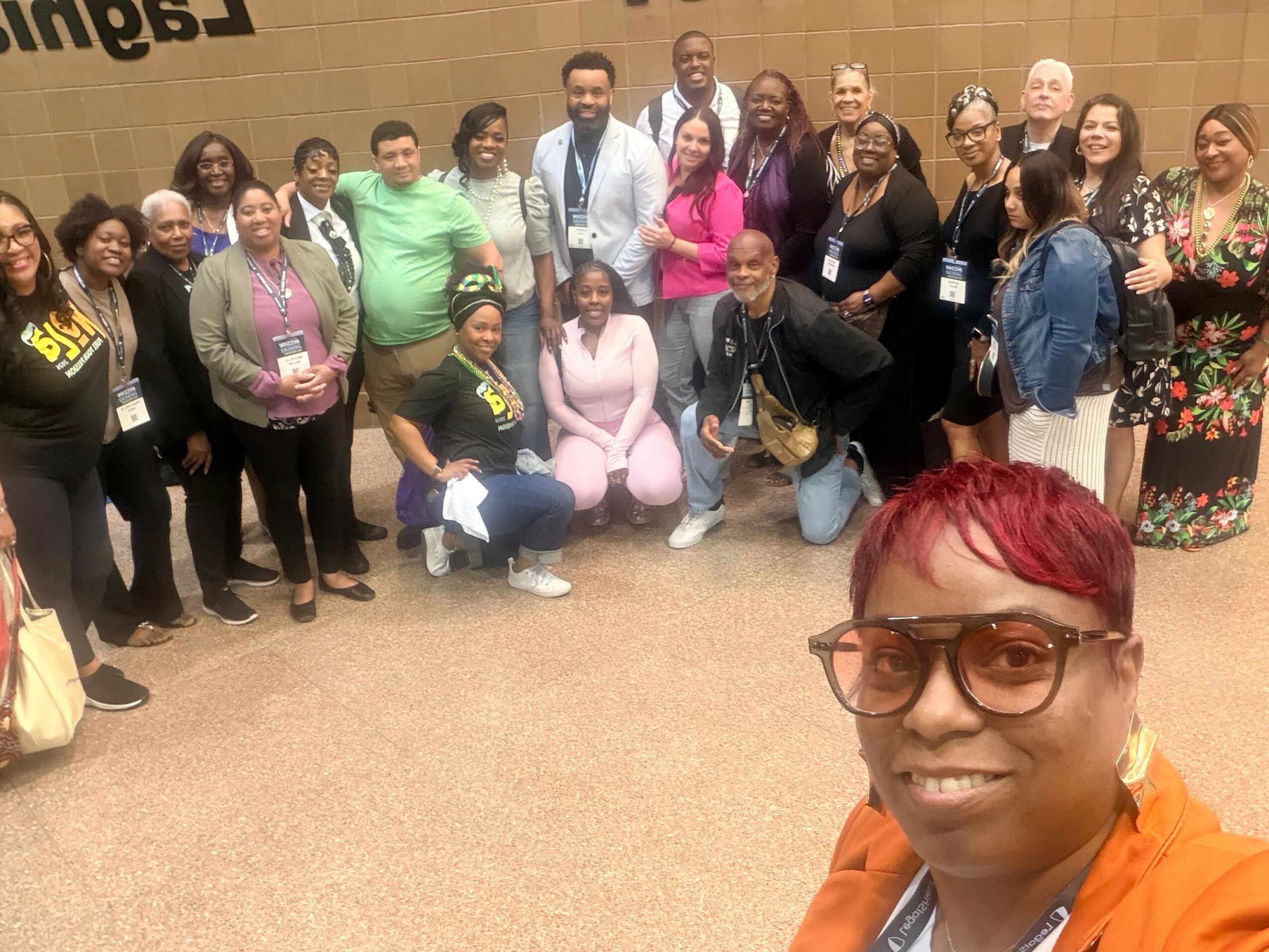 Group of diverse people posing together indoors, some standing and some kneeling, with a woman in the foreground wearing glasses and an orange jacket.