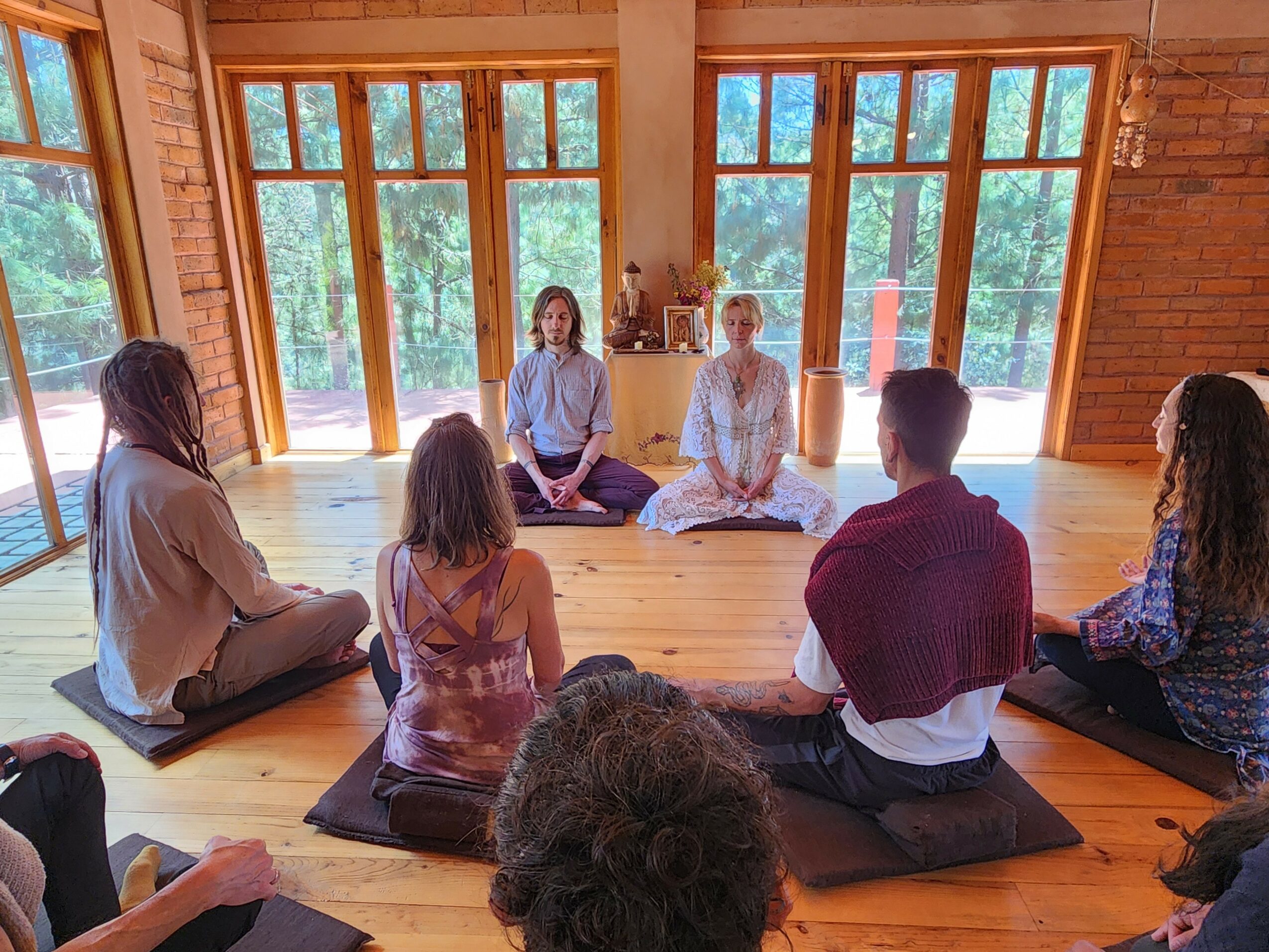 Group of people sitting on mats in a spacious room with large windows and wooden floors, engaging in meditation or yoga.