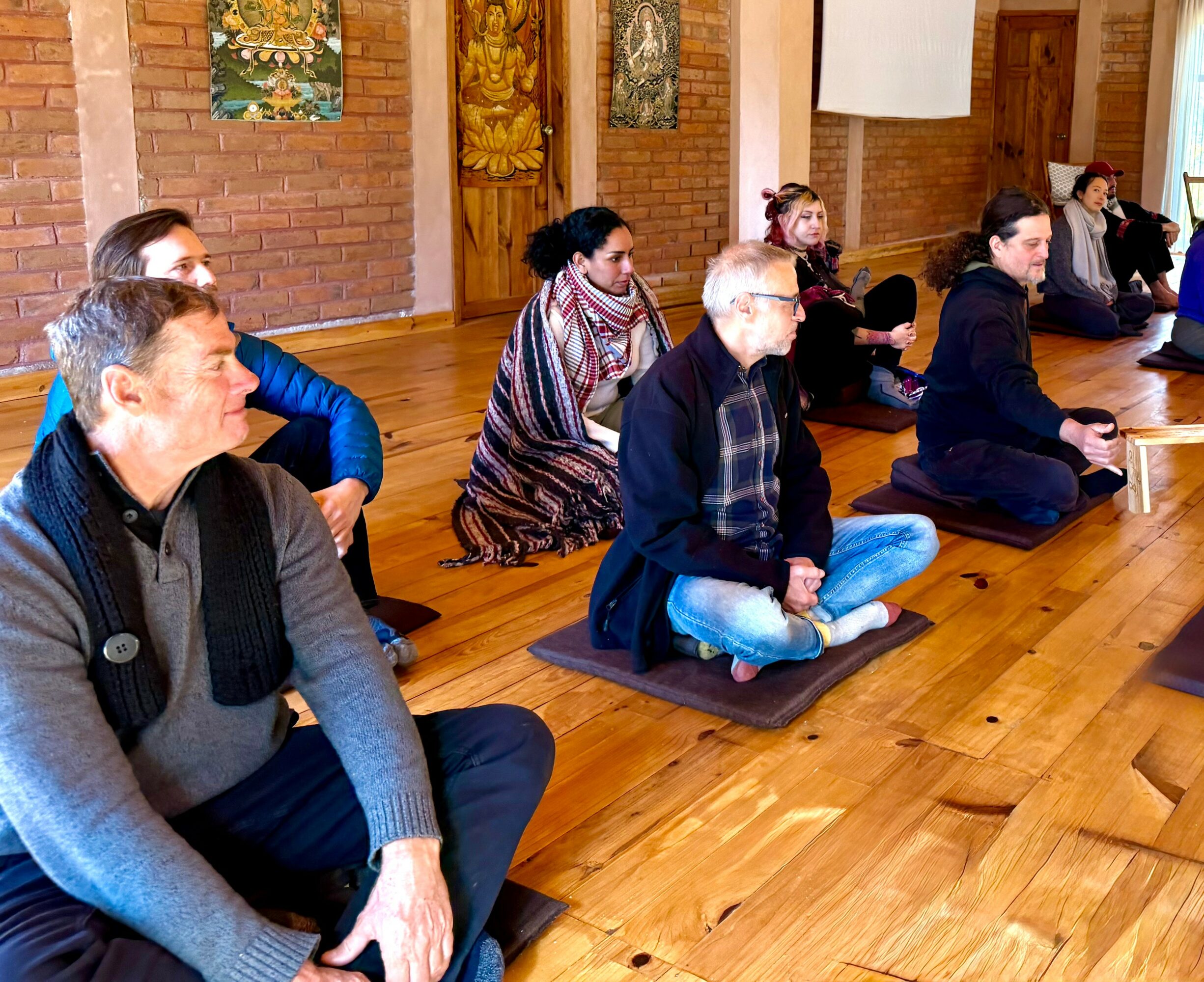 People sitting on mats in a room with wooden floors and brick walls, participating in a meditation or yoga session.