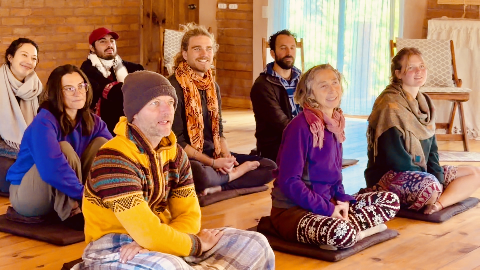 Group of people sitting on the floor in a room with wooden walls, some with eyes closed, meditating or relaxing.