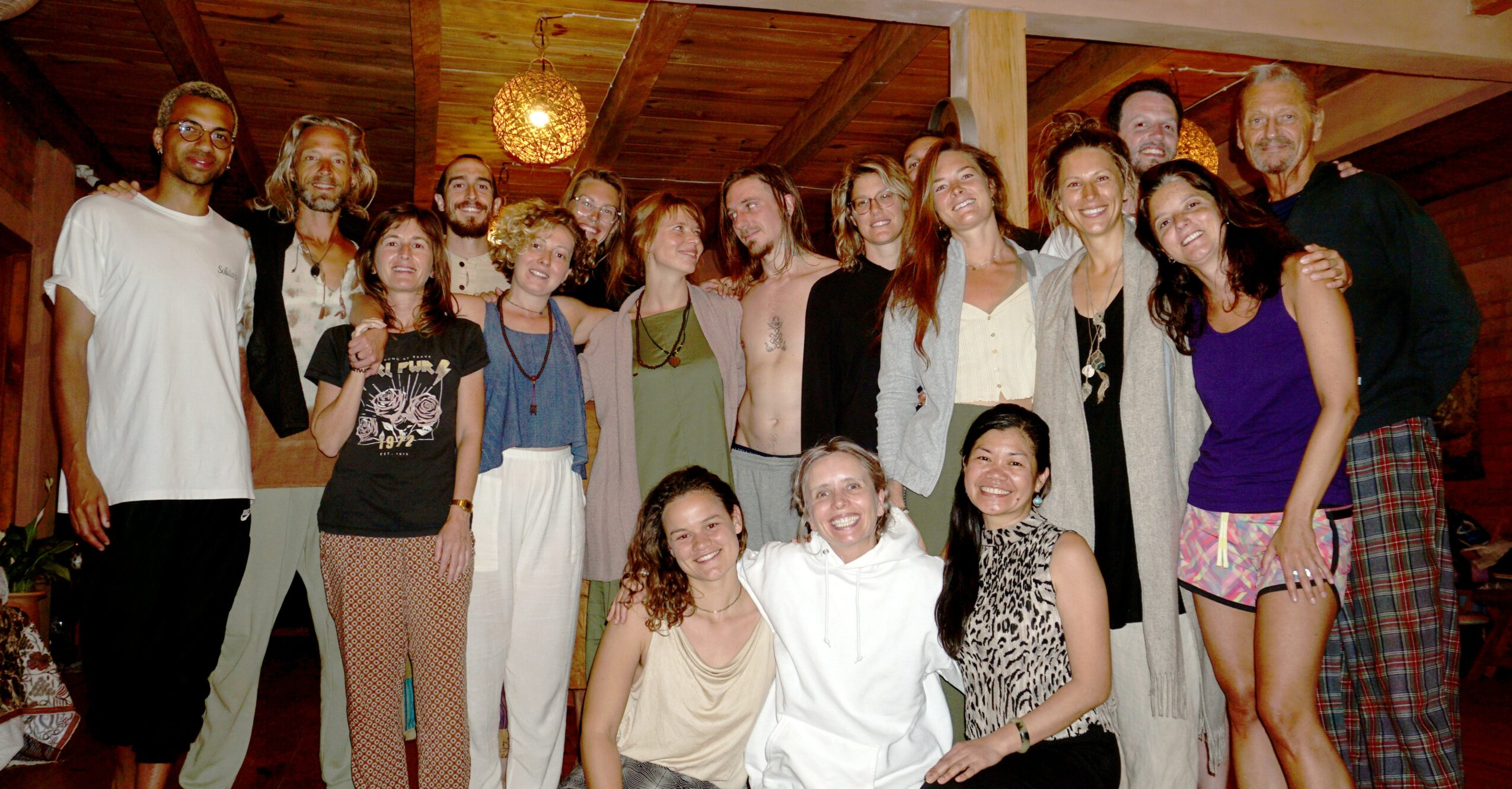 Group of 15 people posing indoors, some smiling, in casual clothing, with wooden ceiling and warm lighting.