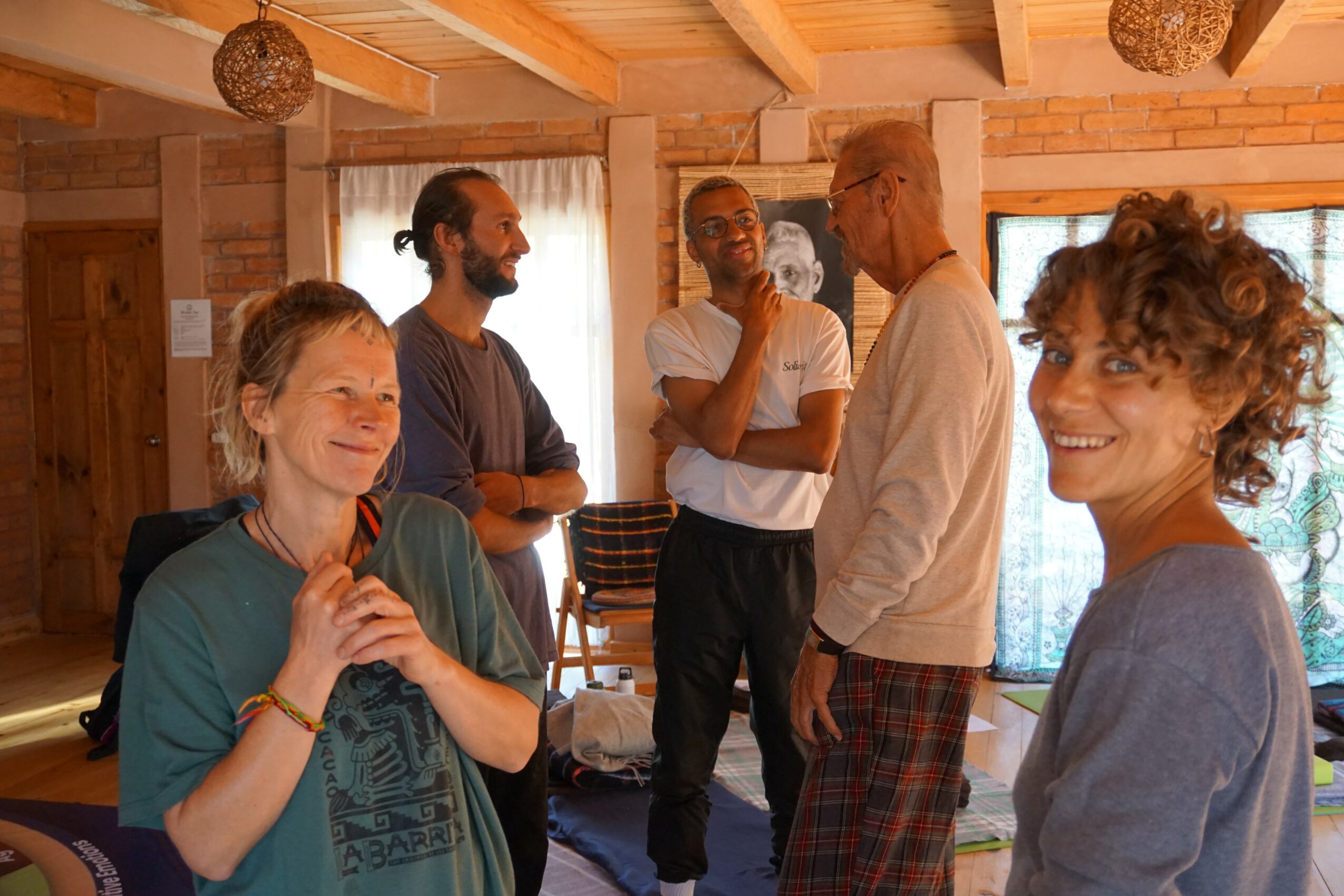 Group of five people smiling and talking indoors with wooden walls and large windows, casual setting.