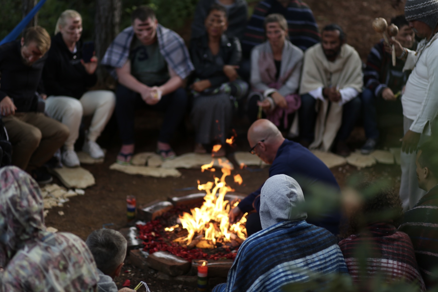 Group of people sitting outdoors around a fire, with a person kneeling near the flames, surrounded by rocks and trees.