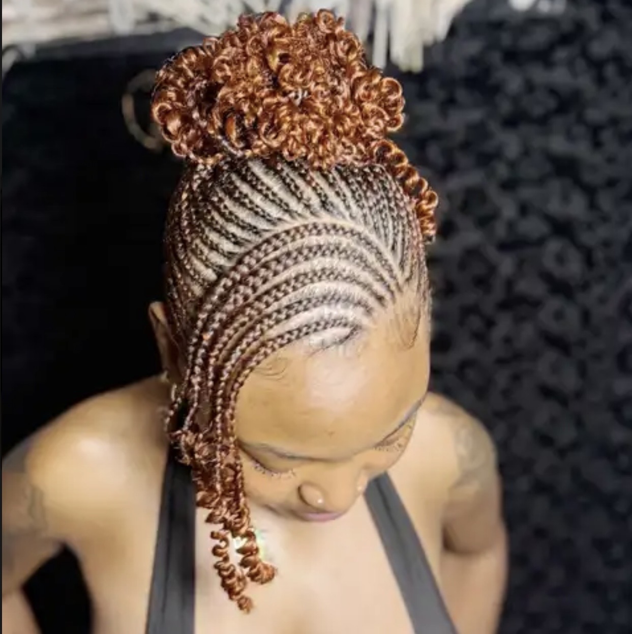 Top view of a woman with braided hair styled in a high bun, with intricate braids on her scalp, against a dark background.