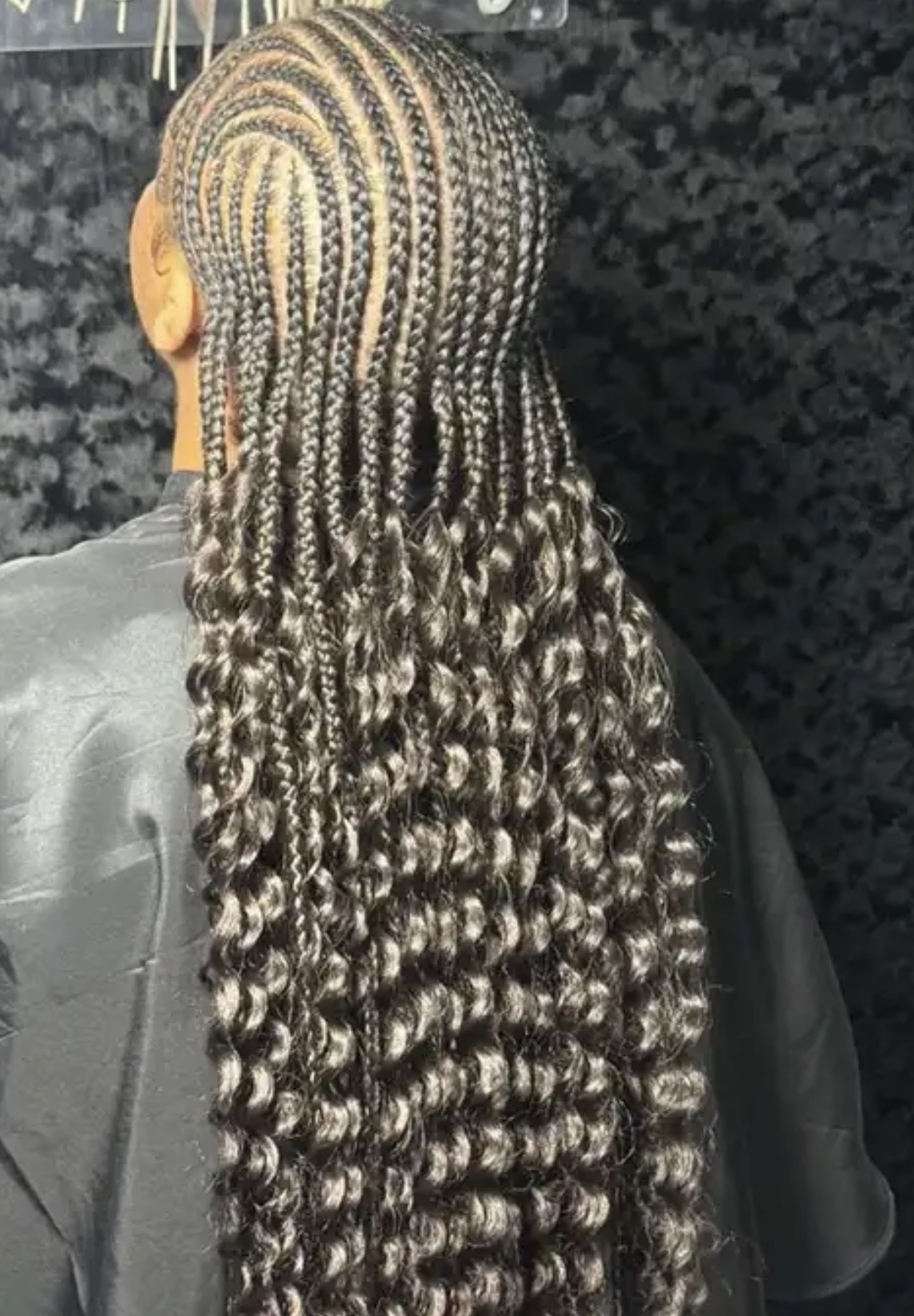 Person with intricately braided hair styled in tight curls, seen from the back, with a dark background.